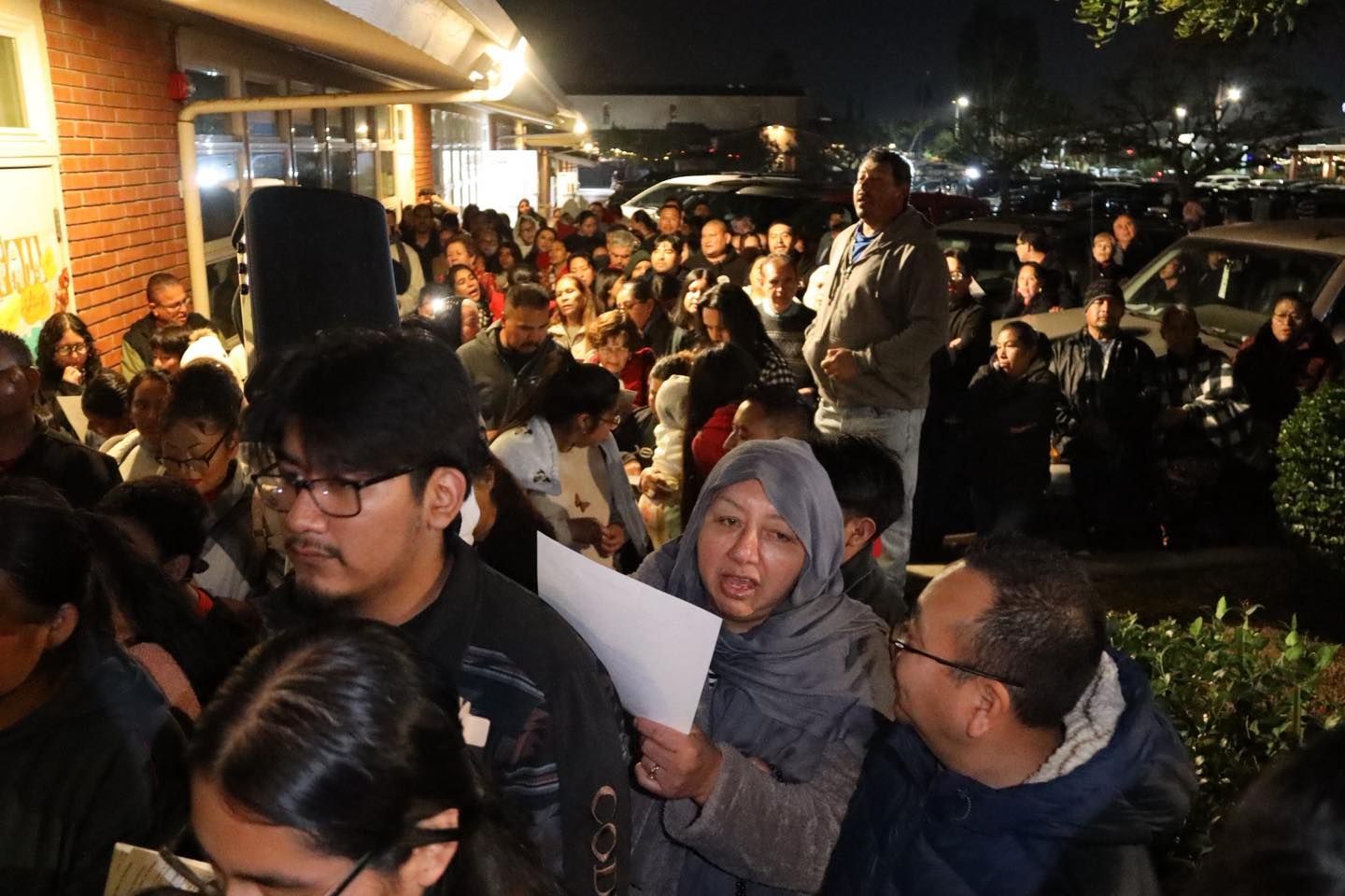 Crowded nighttime gathering outside a building, with people seated and standing under warm lights