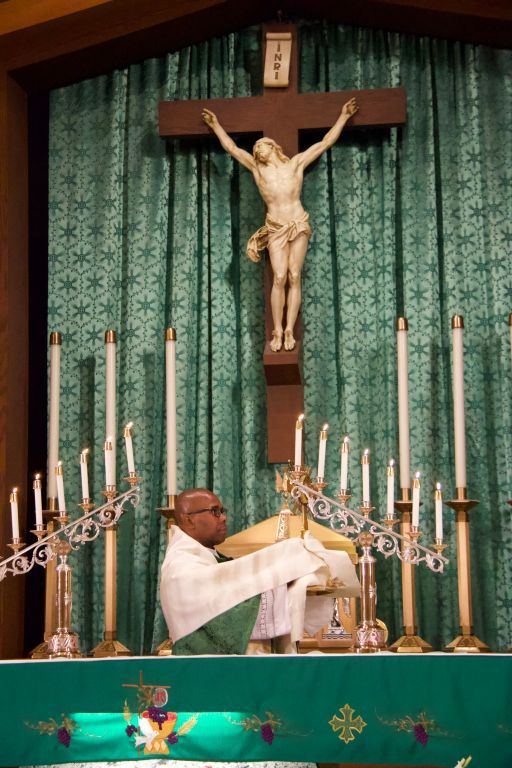 Priest elevates the Eucharist at an altar beneath a crucifix, surrounded by candles and green drapery.