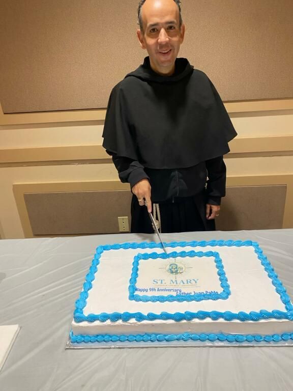Man standing behind a blue-and-white frosted cake on a table, smiling in a banquet room.