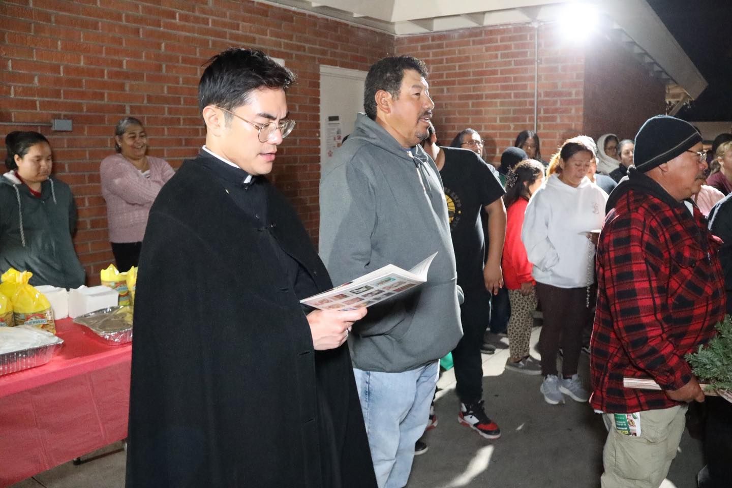 People gathered indoors; a man in a black coat reads from papers near a table with red cloth.