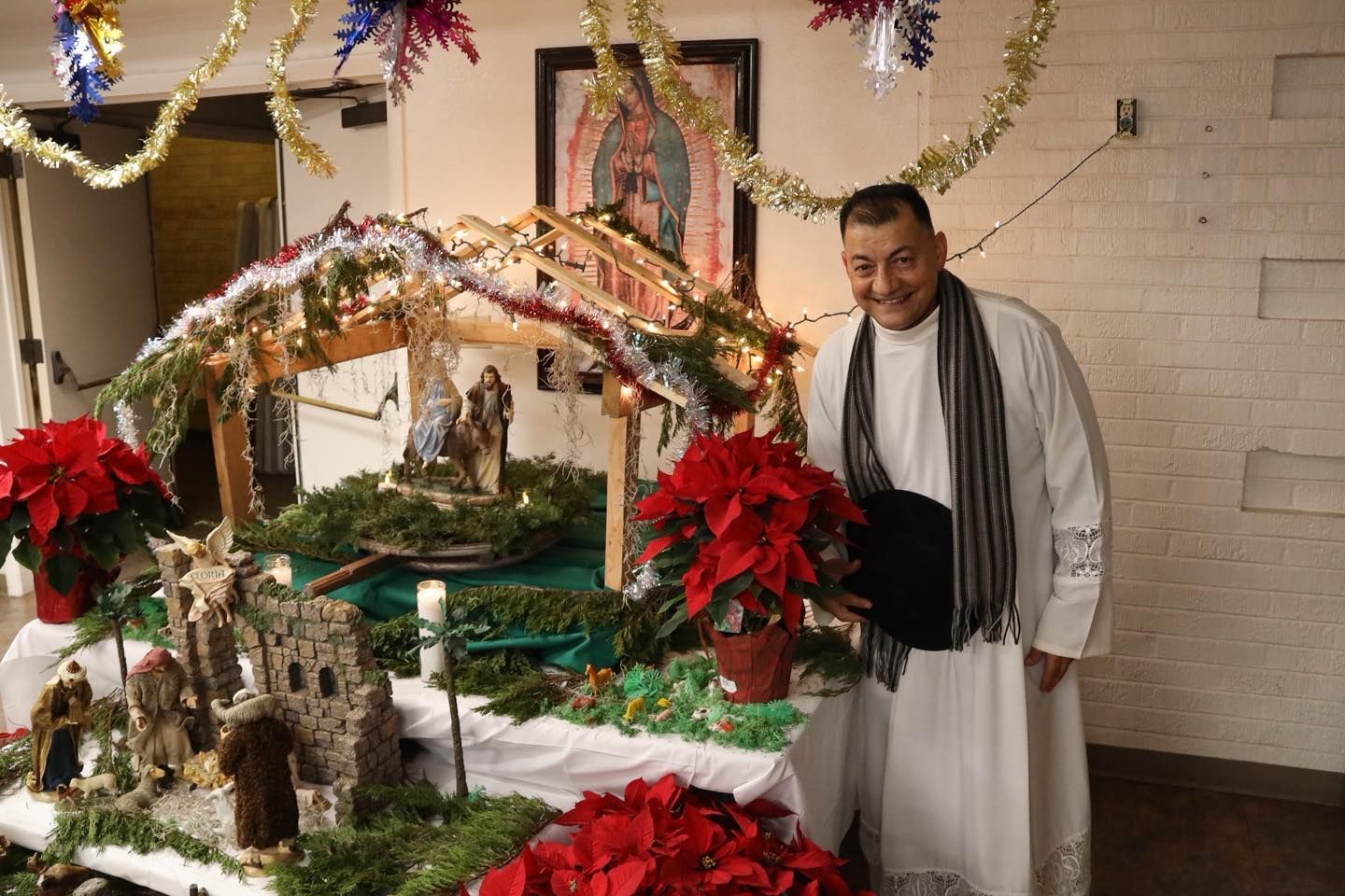 Man standing beside a decorated nativity scene with red poinsettias indoors