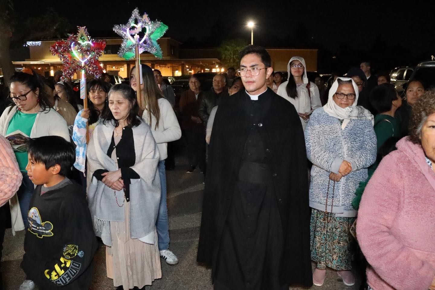 Crowd gathered outdoors at night, many holding colorful star lanterns, with a man in a black coat in front