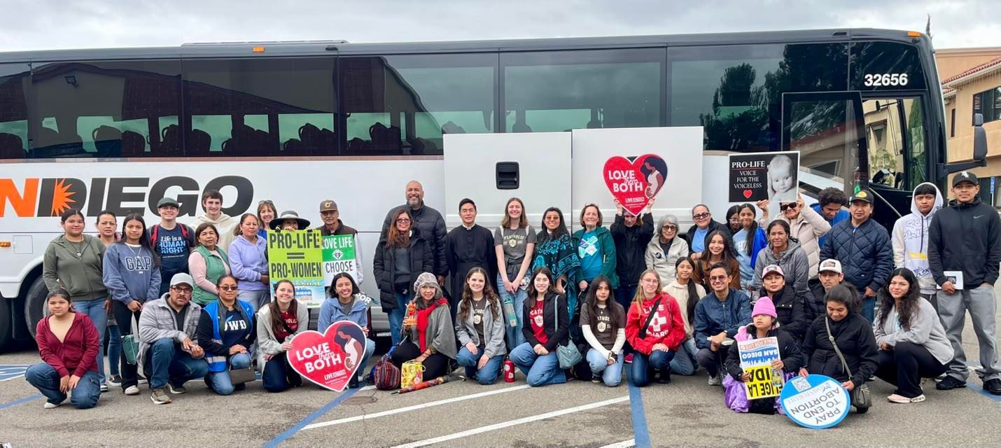 Large group posing outside a black tour bus, holding signs and smiling in a parking lot.