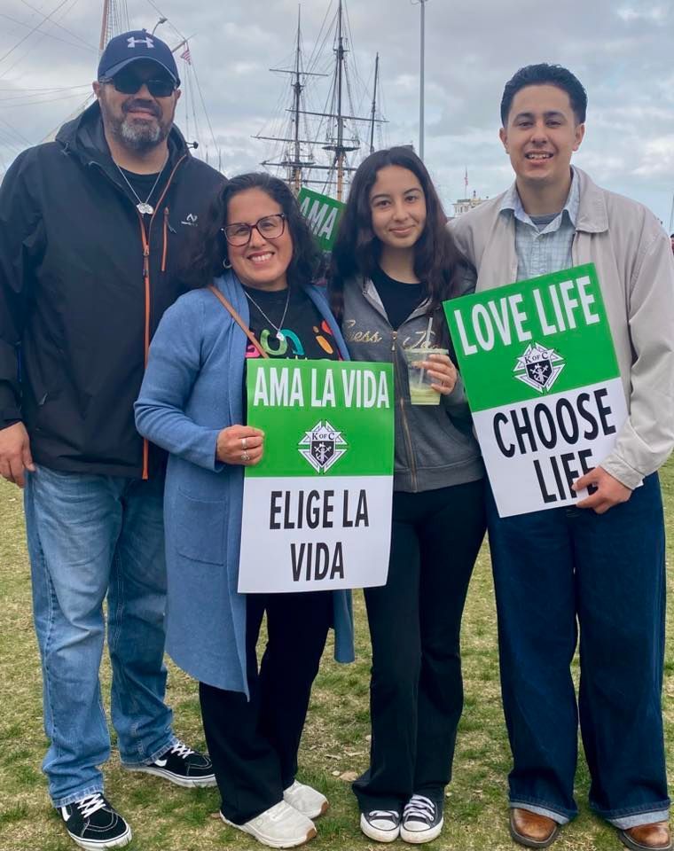 Four people outdoors holding pro-life signs in front of a sailing ship.