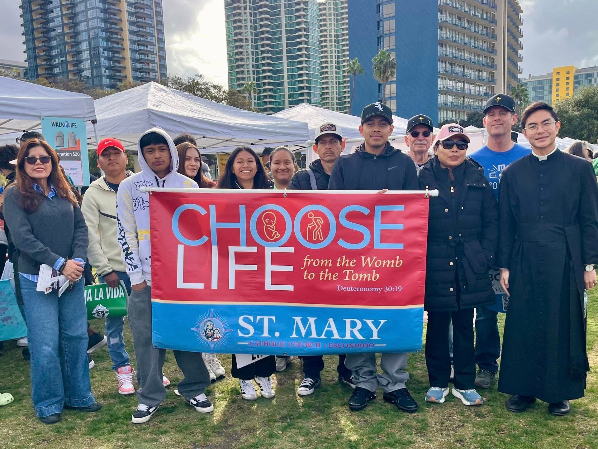 Group holding a red “Choose Life” St. Mary banner at an outdoor event with tents and city buildings behind them