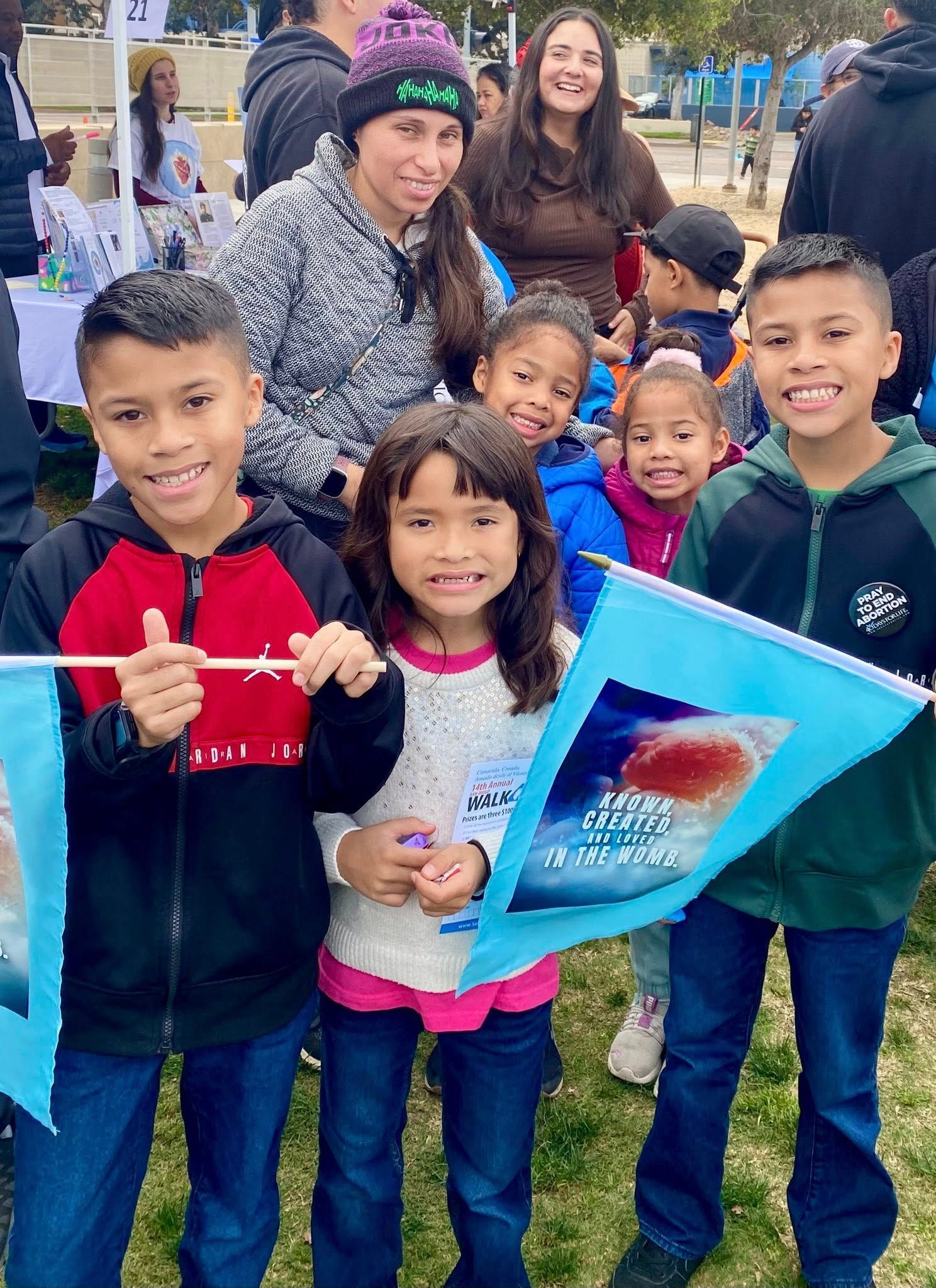 Children posing outdoors at a winter event, smiling and holding a red toy and blue folder.
