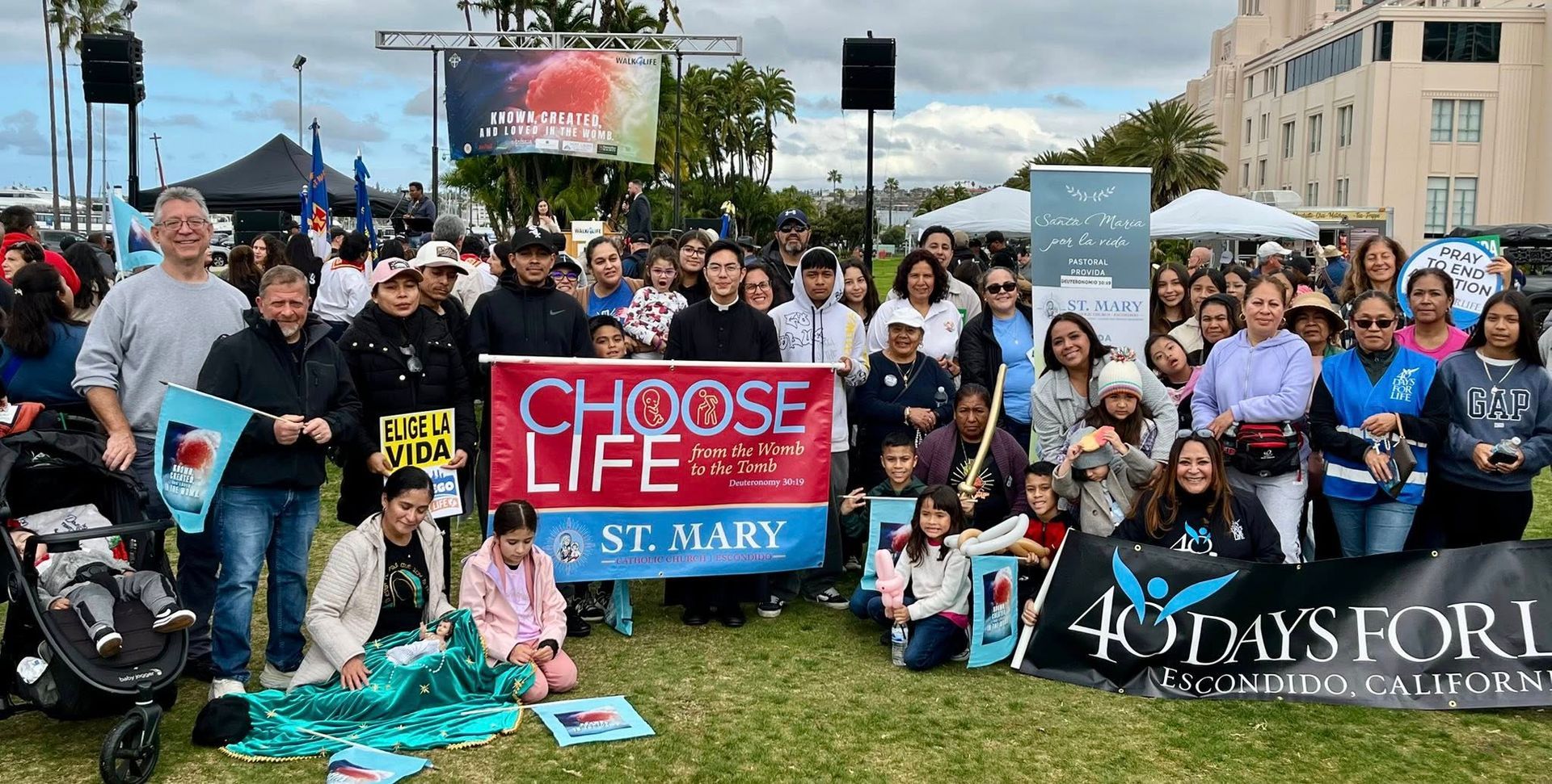 Crowd holding “Choose Life” and “40 Days for Life” signs at an outdoor rally, with a crowd and city backdrop