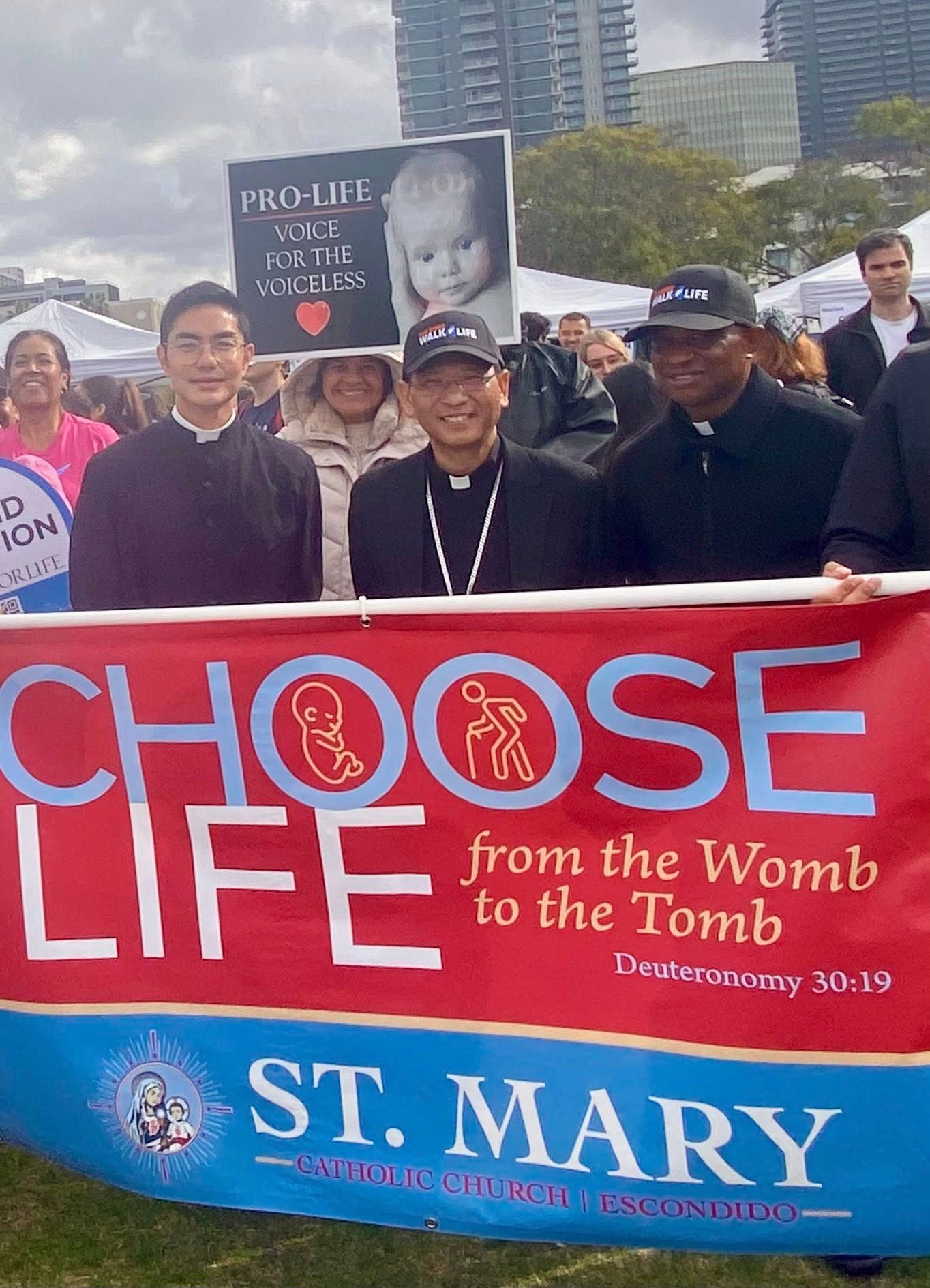 People holding a red banner reading “Choose Life from the Womb to the Tomb” at a St. Mary event
