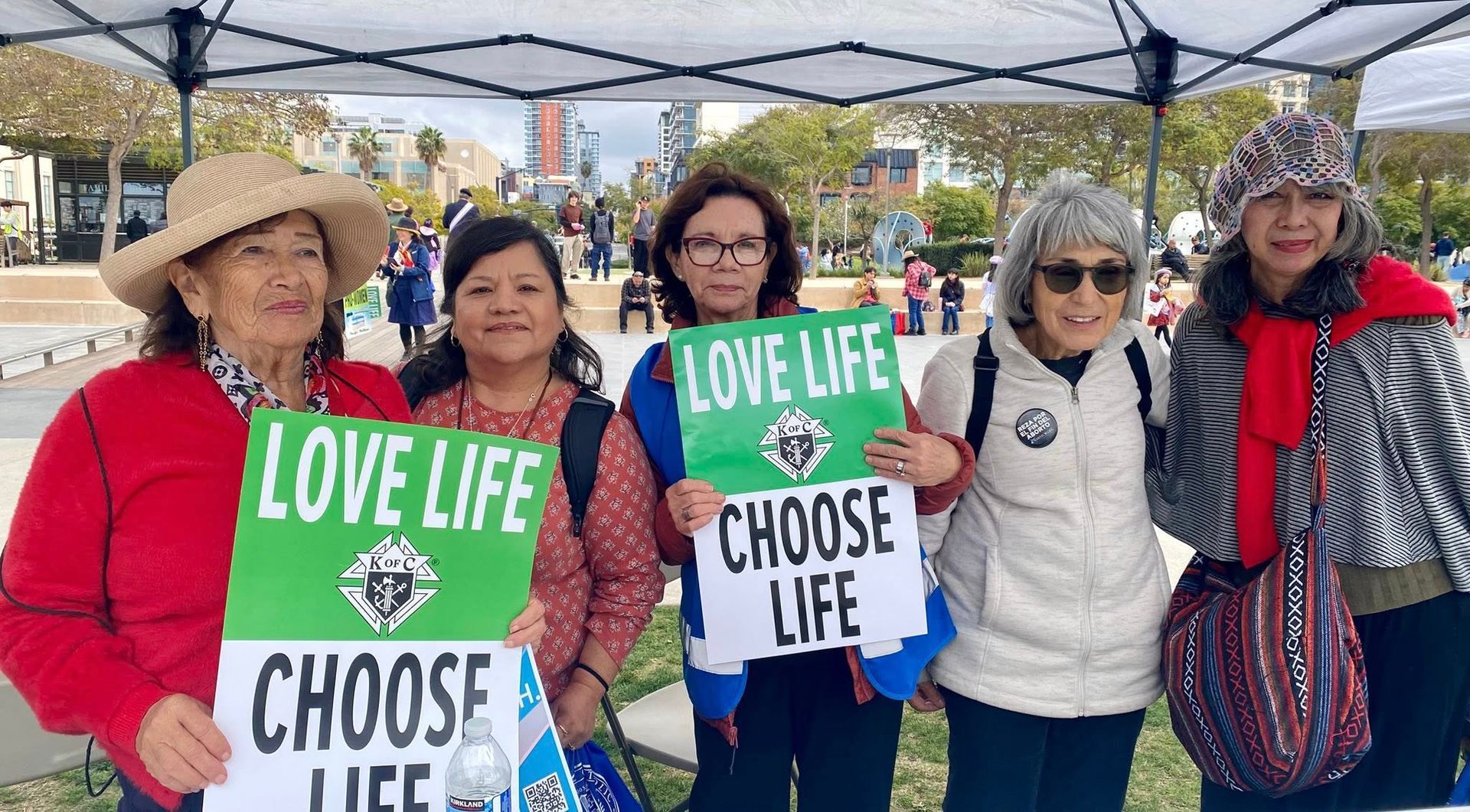 Five people at an outdoor event hold “LOVE LIFE CHOOSE LIFE” signs under a canopy.