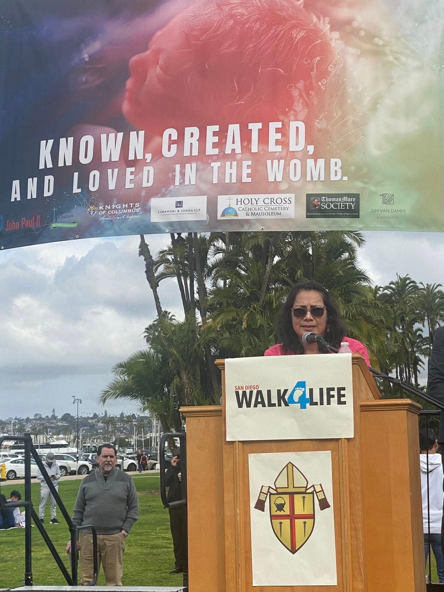 Woman speaking at Walk4Life podium beneath “Knitted, created, and loved in the womb” banner outdoors.