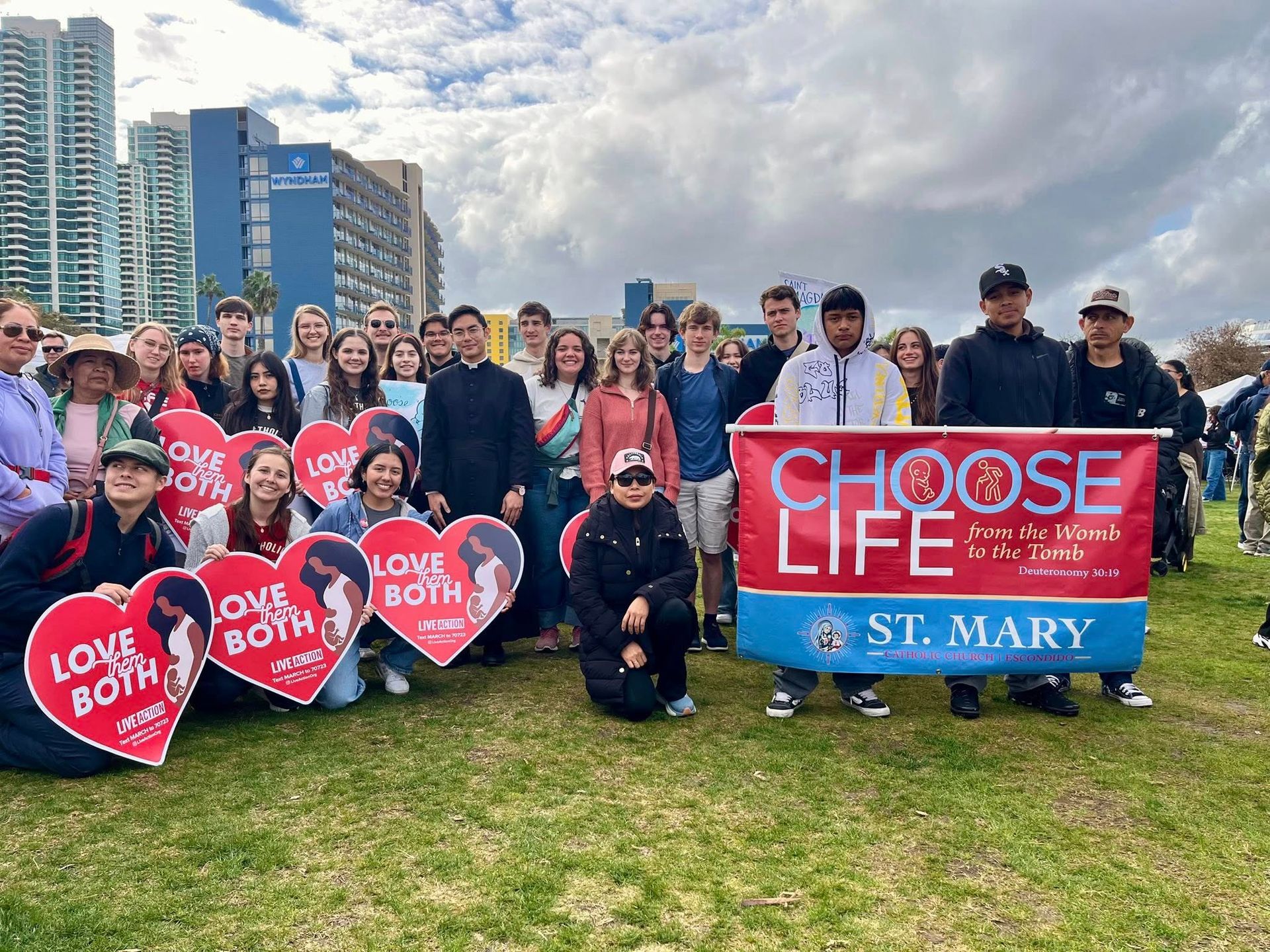 Group posing on a grassy field holding red “Love Both” and blue “Choose Life” signs at a St. Mary event