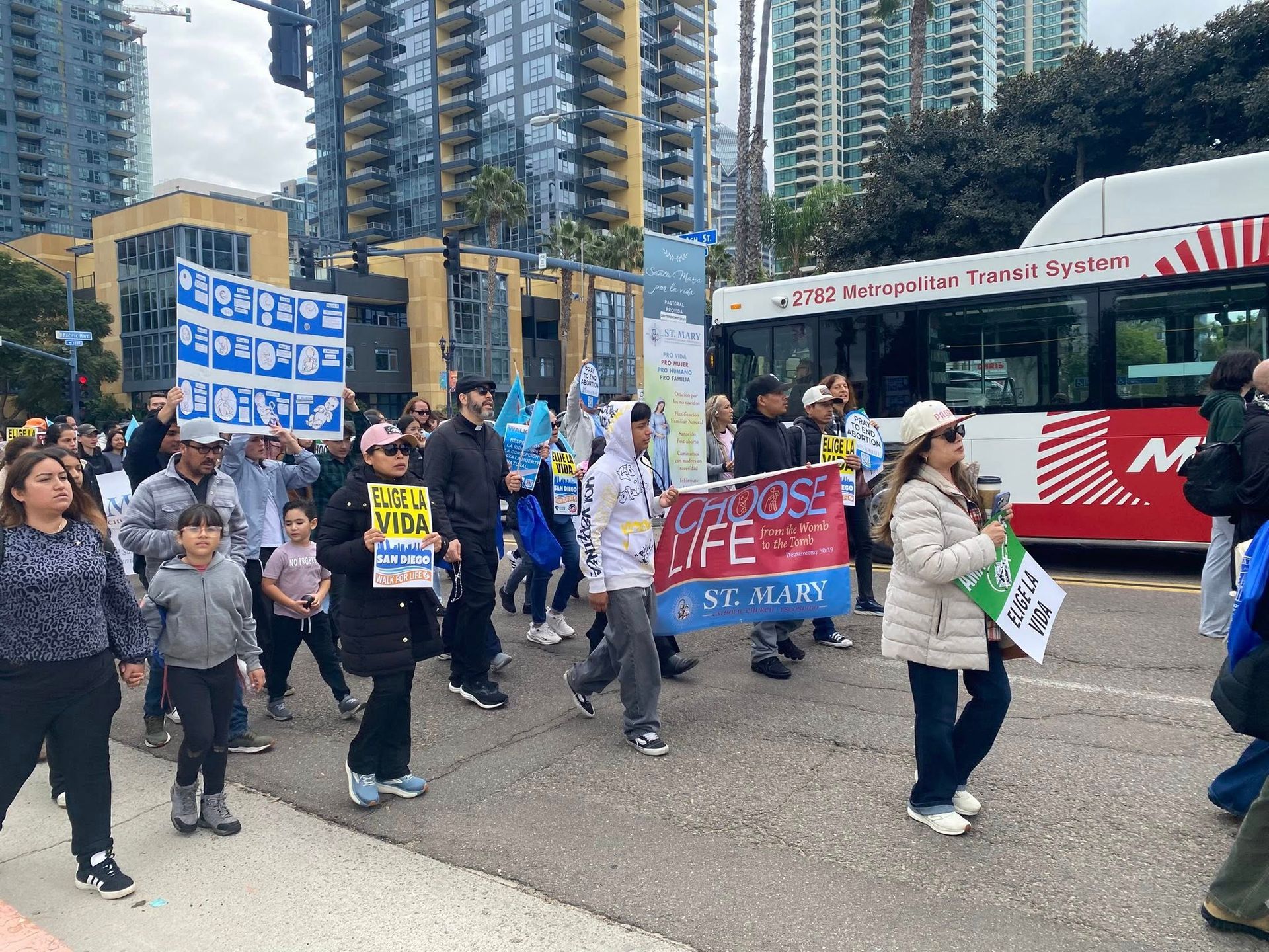 Protest march with people holding signs and banners near a red-and-white bus in a city street