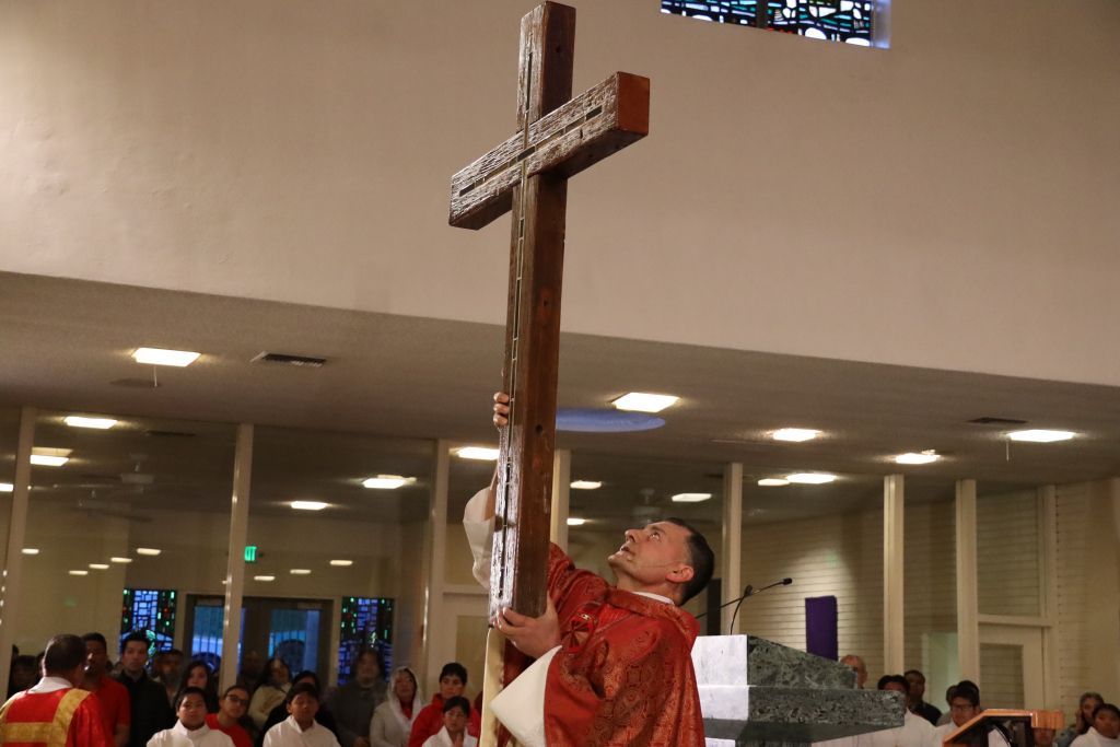 Person in red robe raising a large wooden cross indoors during a church ceremony