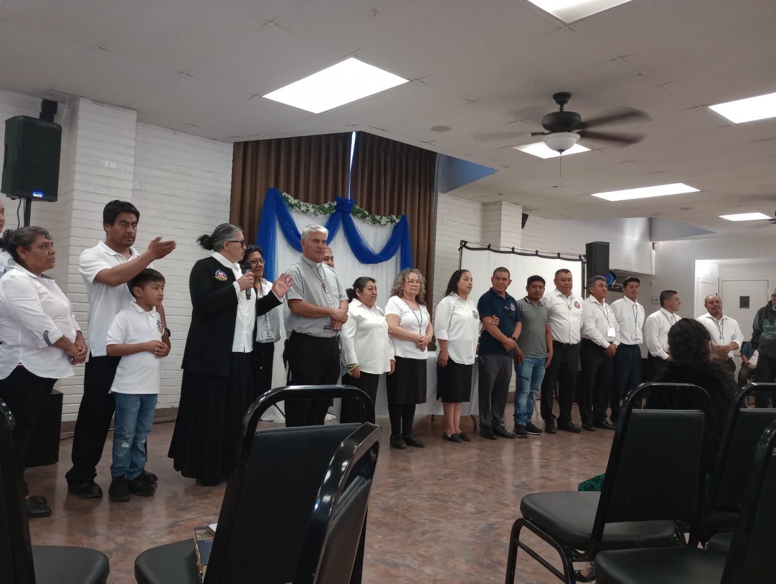 People standing in a line inside a hall, singing or reciting beside a decorated backdrop.