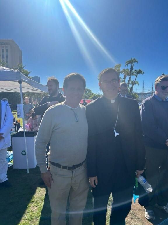 Two people standing outdoors in sunlight at a social event, with a crowd and blue sky behind them.