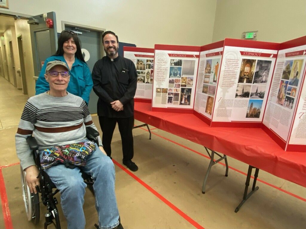 Three people at a display booth with red-draped tables and poster boards, one seated in a wheelchair.
