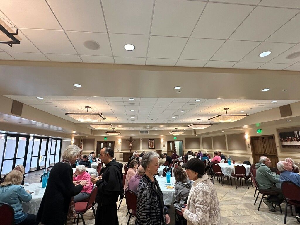 People seated at round tables in a brightly lit banquet hall with a dropped ceiling and large windows.
