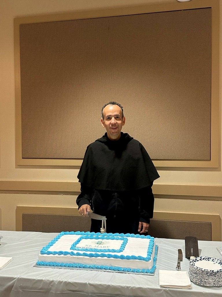 Man standing behind a table with a blue-and-white cake in a room with a corkboard backdrop