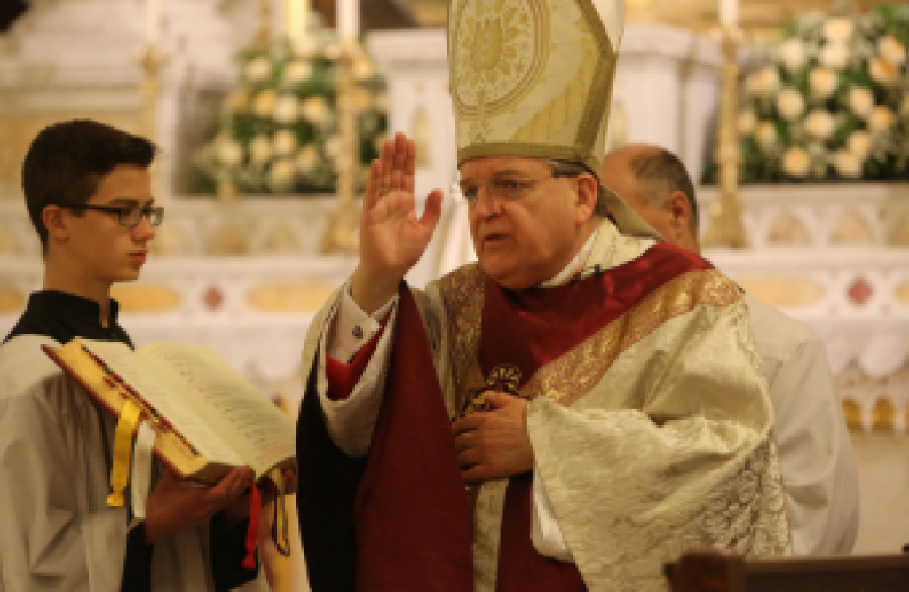 Catholic bishop in gold vestments raises a hand during a church service beside an altar server.
