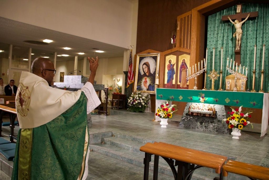 Priest in green vestments reads at altar in a church, with crucifix, candles, and floral arrangements.
