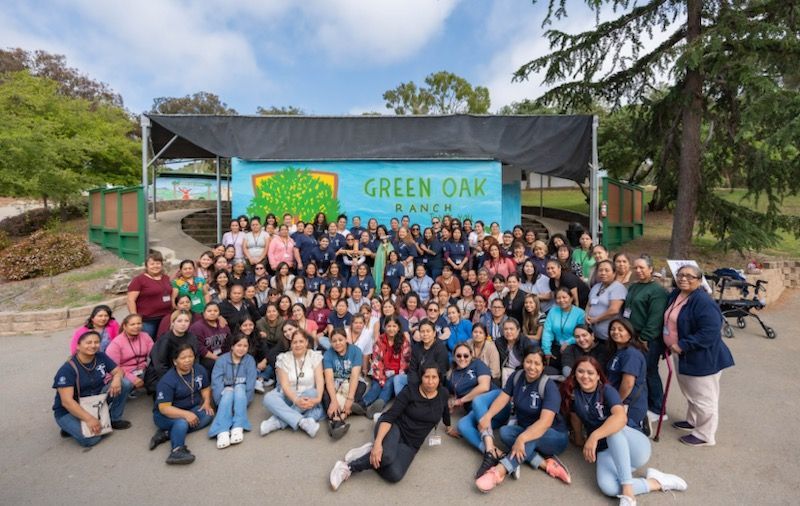 Large group posing outdoors in front of a green 