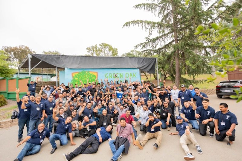 Large group posing outdoors in front of a building, many in navy shirts, some seated on the ground.