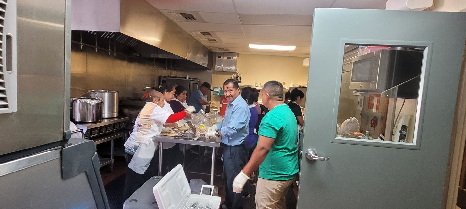 Workers in a commercial kitchen serving food at a counter, with stainless steel appliances and a service window.