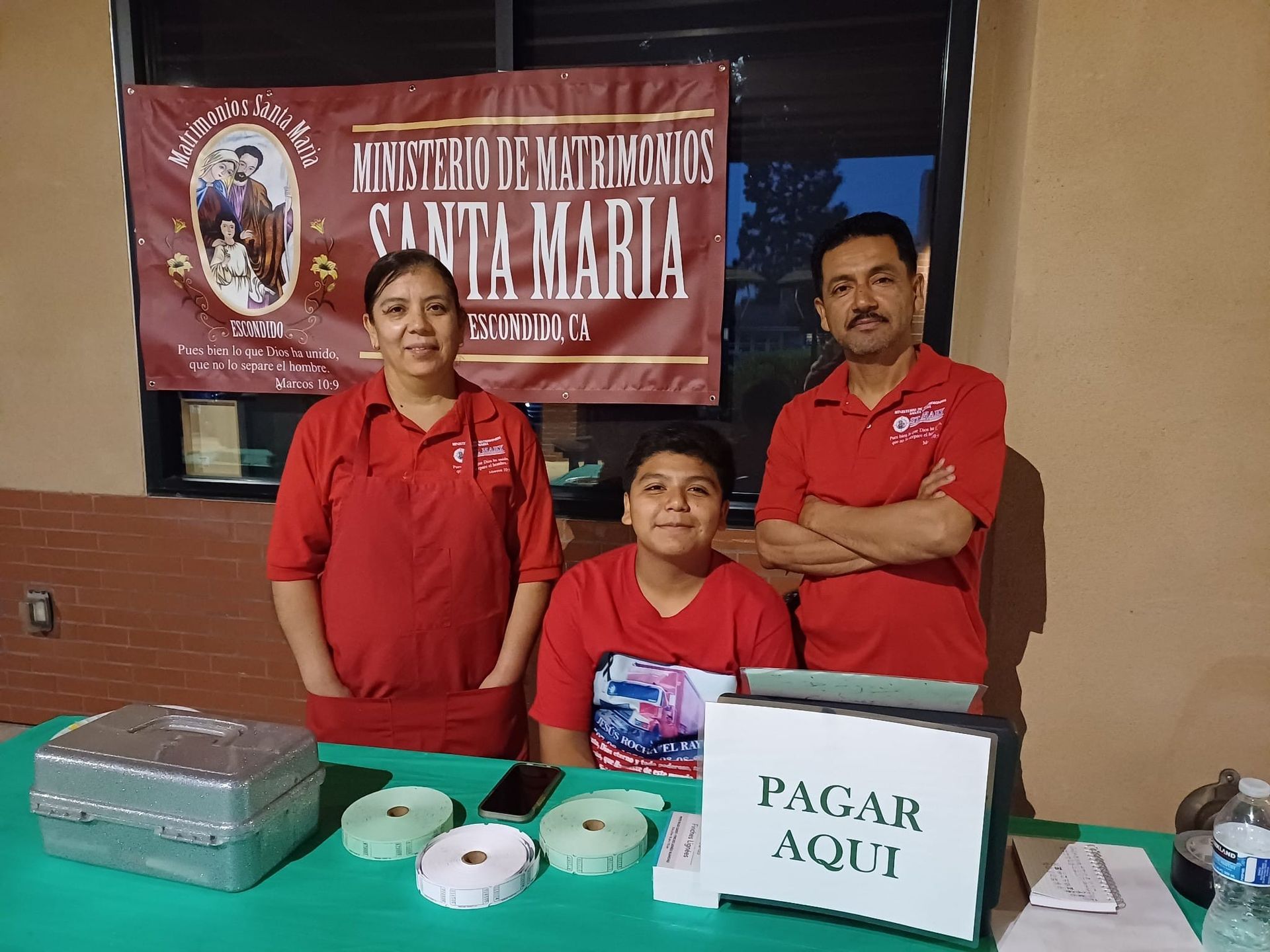 Three people behind a table at a school event, with a donation box labeled “PASAR AGUI” and a banner in back.