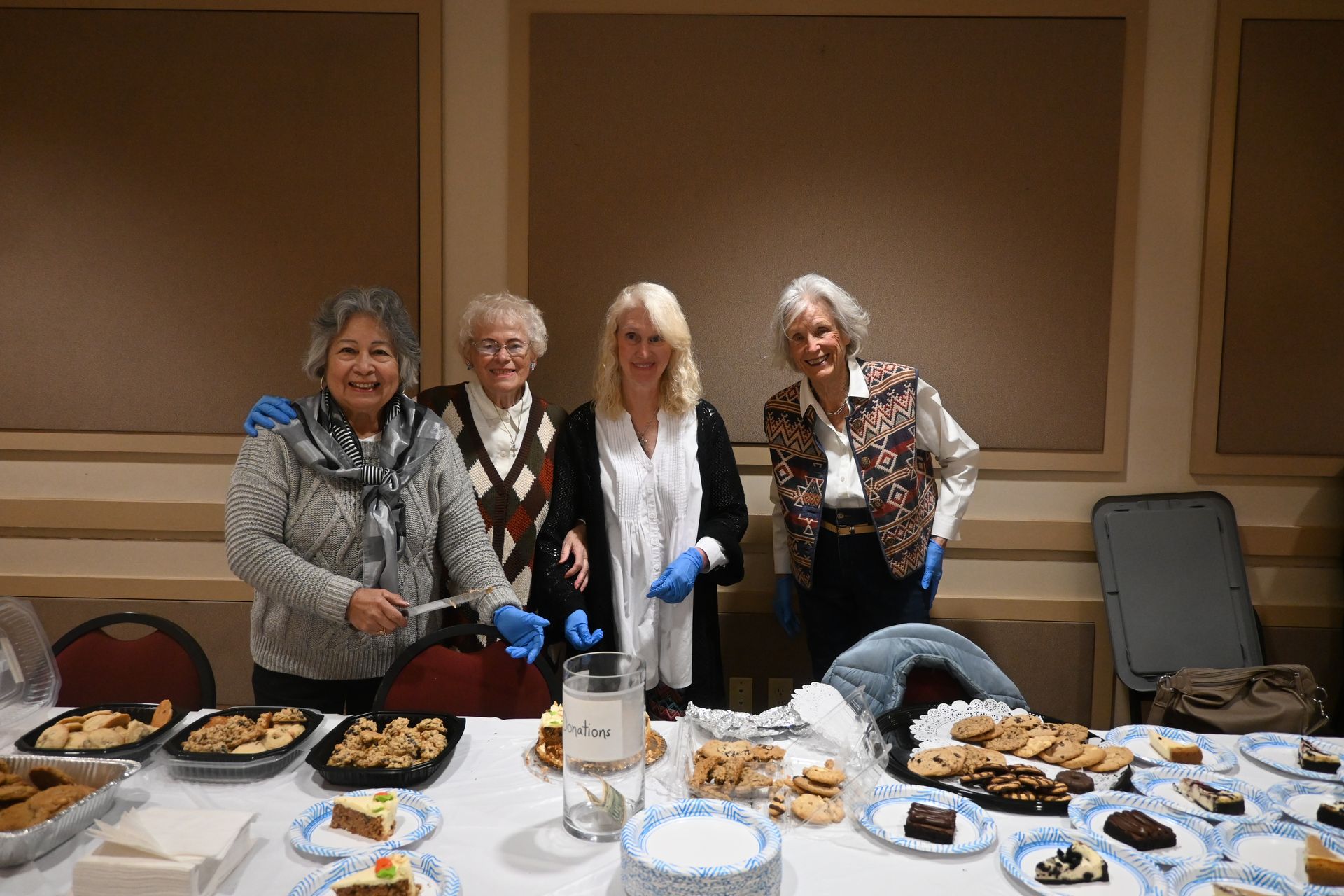 Four people standing behind a table of assorted baked goods in a room, posing for a photo.