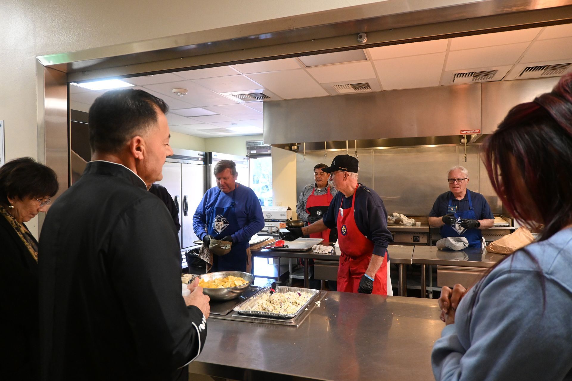 People gathered around a stainless-steel buffet counter in a kitchen or cafeteria.