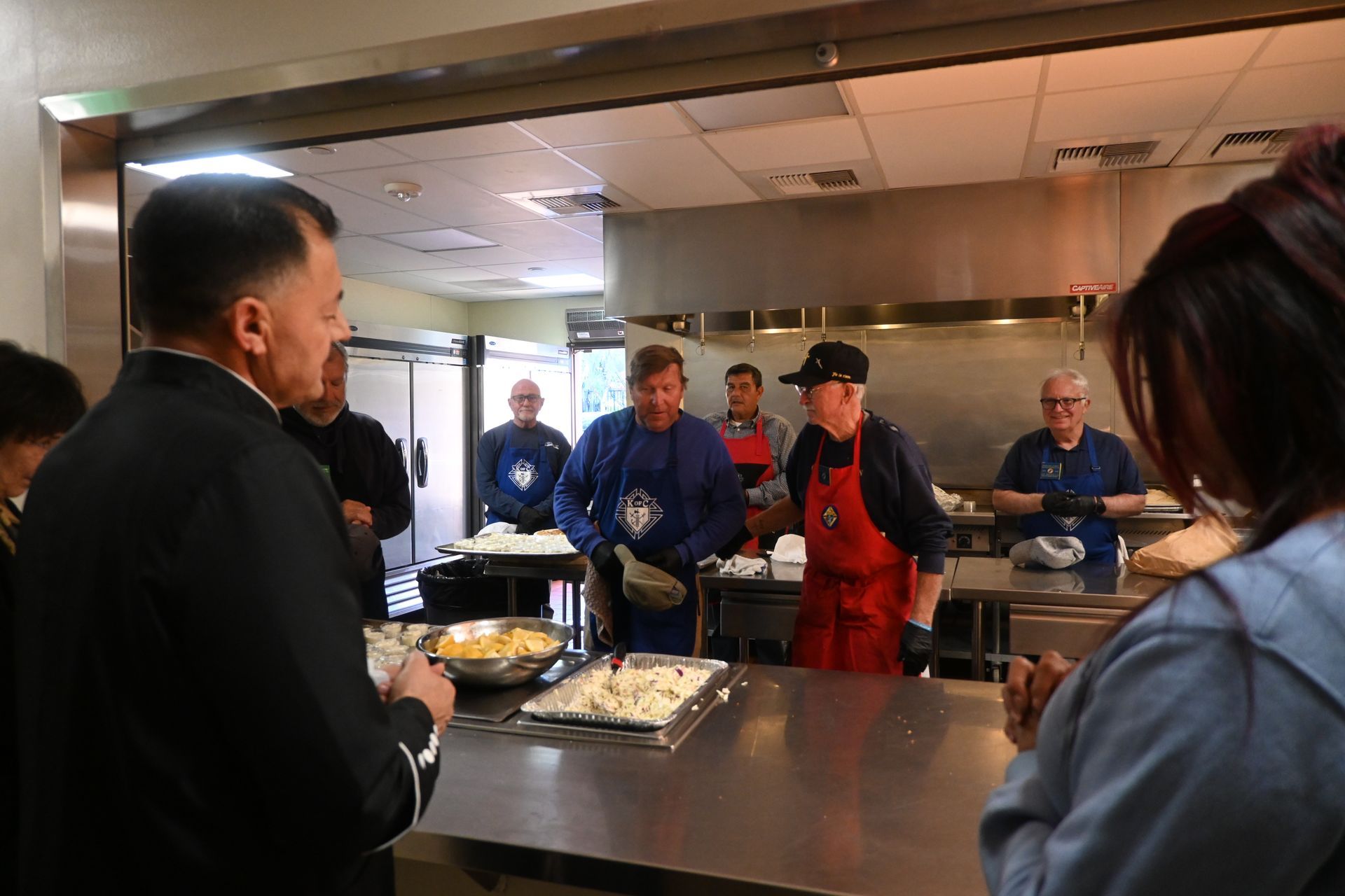 People standing at a cafeteria counter receiving food from staff in a stainless-steel serving area.
