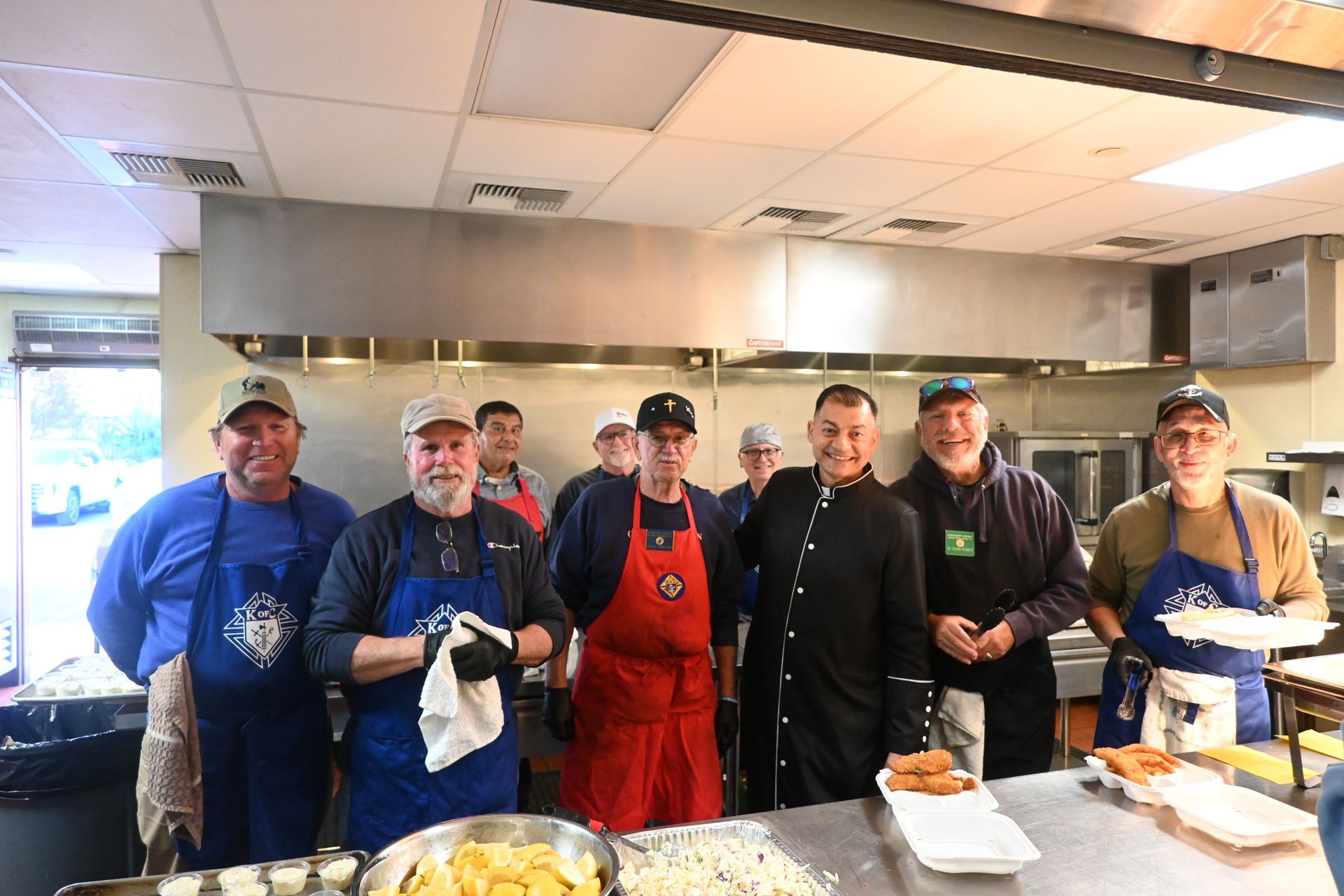 Group of volunteers in a kitchen, standing behind a counter with prepared food and trays.