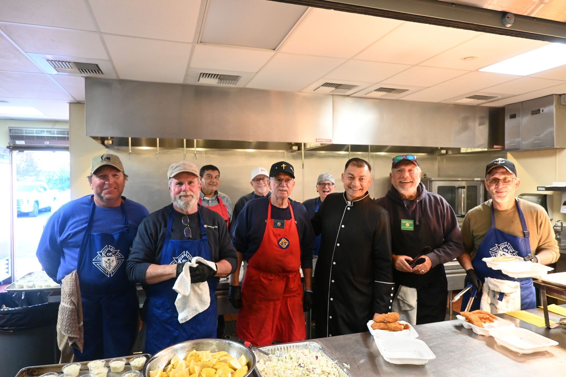 Volunteers in an industrial kitchen smiling behind a counter with food trays and utensils