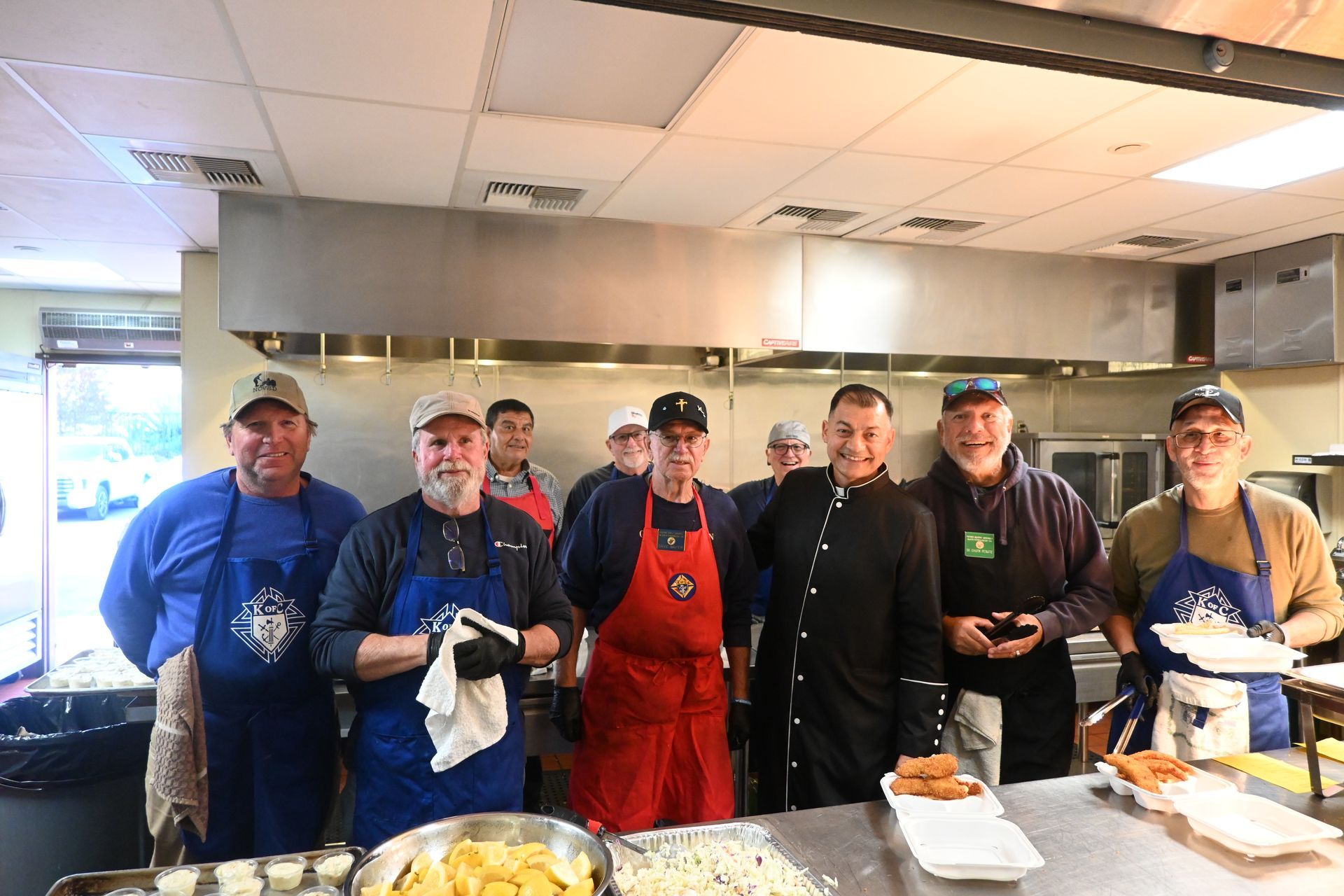 Group of cooks standing behind a buffet counter in a kitchen, wearing aprons and hats.