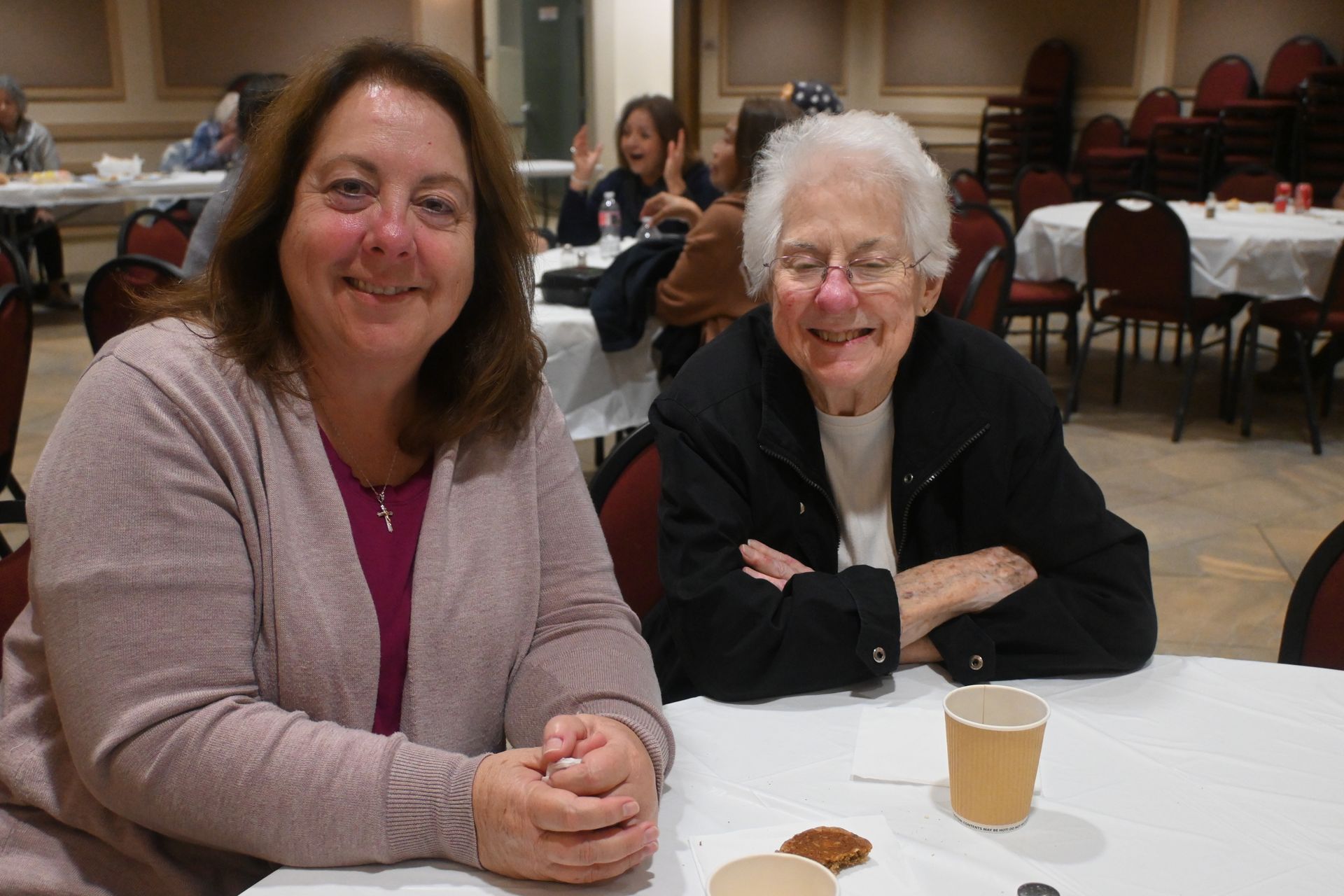 Two women sit at a round table in a dining hall, smiling with coffee cups in front of them.