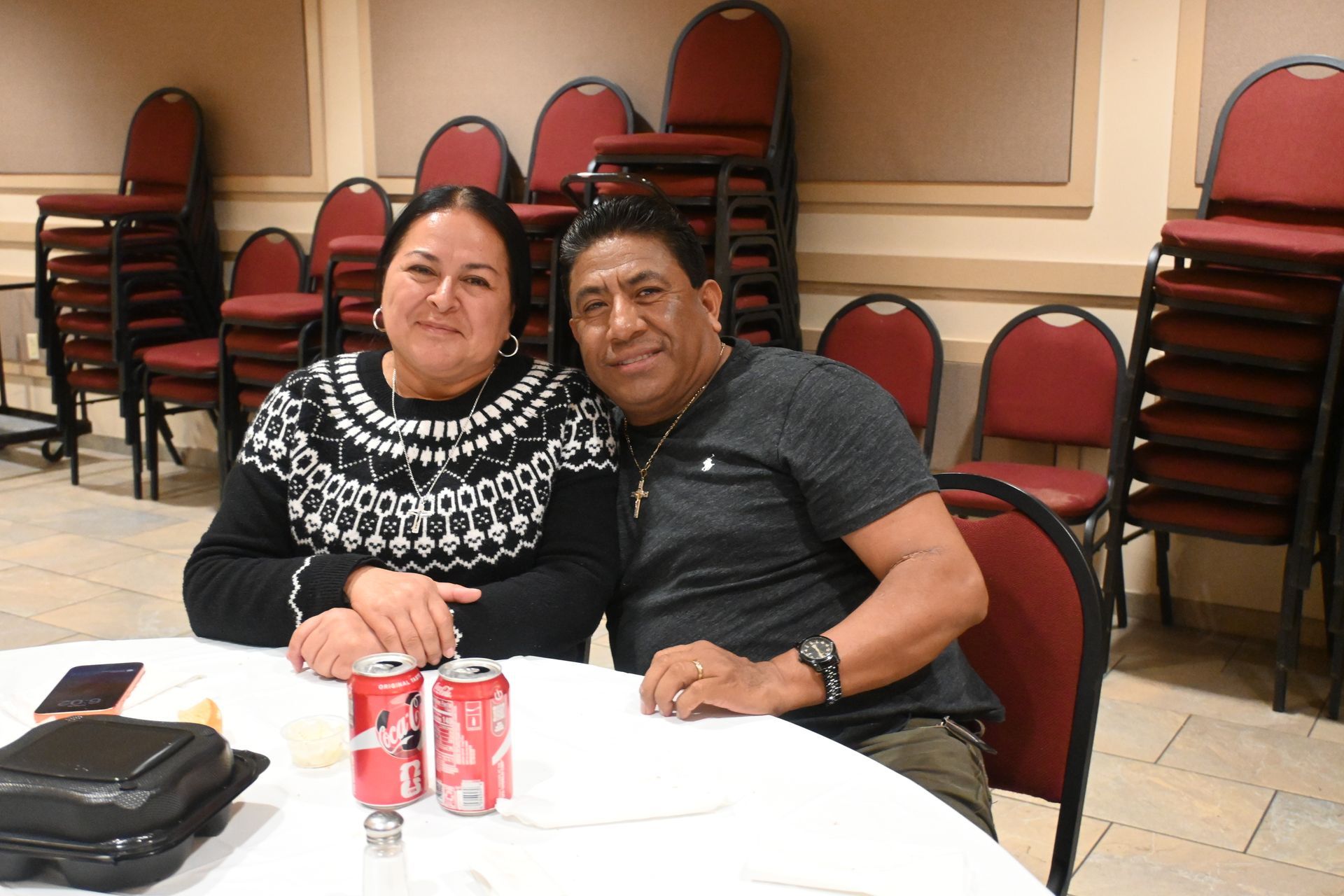 Two people sitting at a table in a banquet room, smiling with soda cans and stacked chairs behind them.