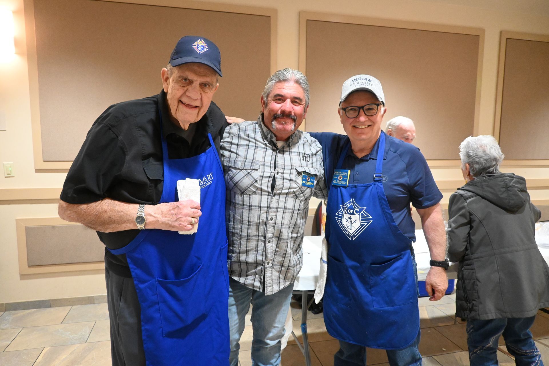Three men in blue aprons and caps smiling indoors at an event