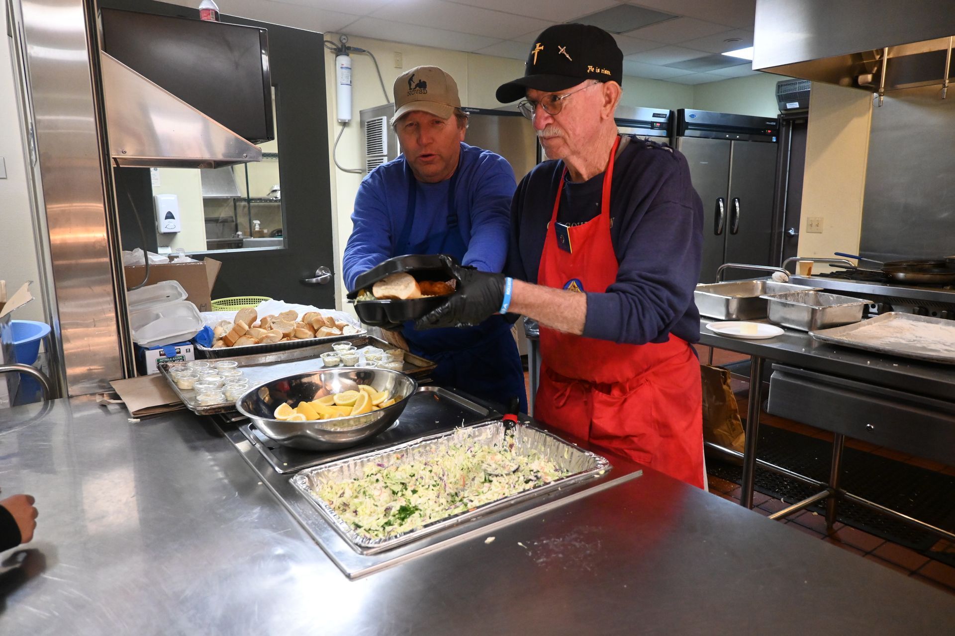 Two workers serving food from stainless-steel trays in a commercial kitchen
