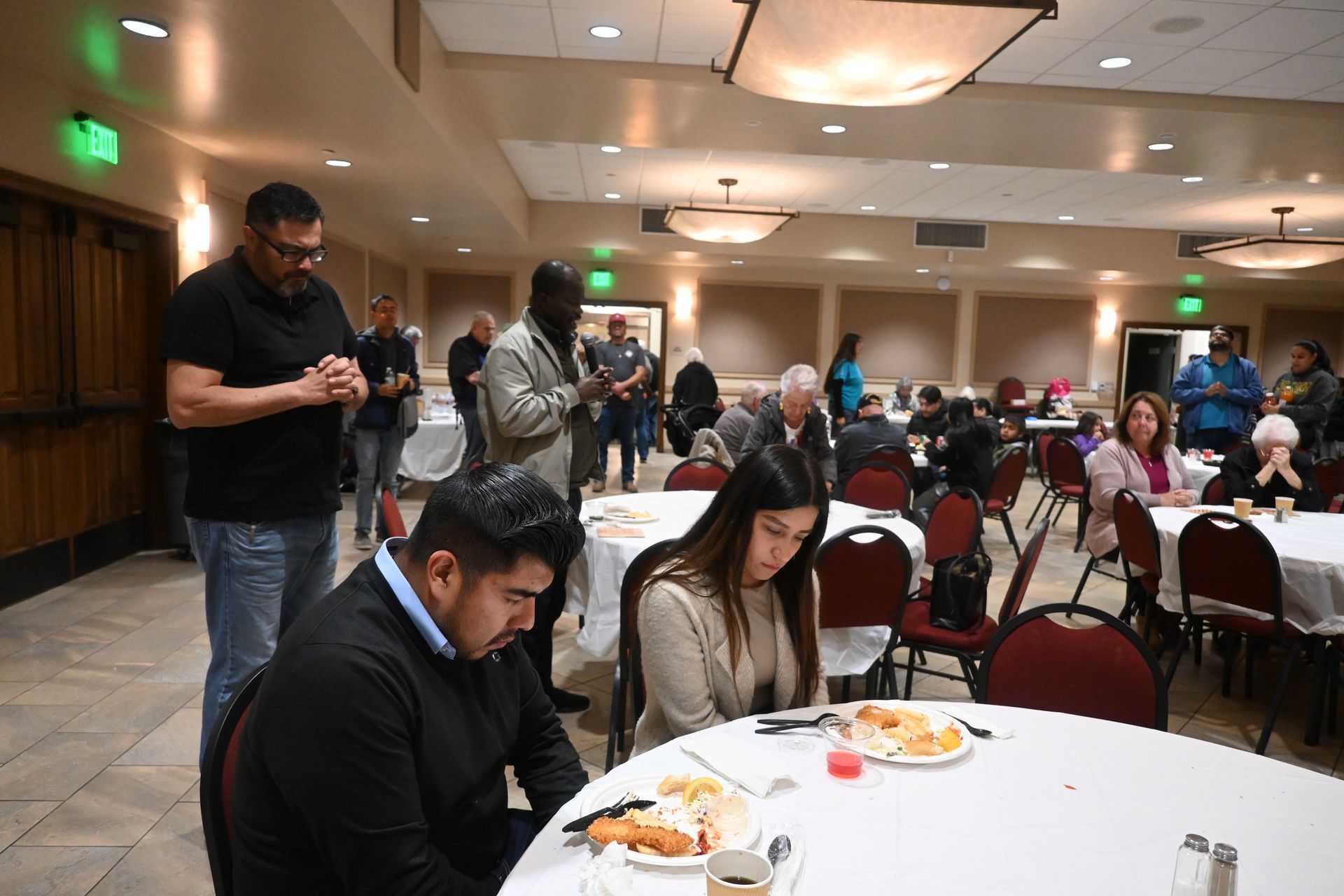 People seated at round tables in a banquet hall, with others standing and eating in the background.