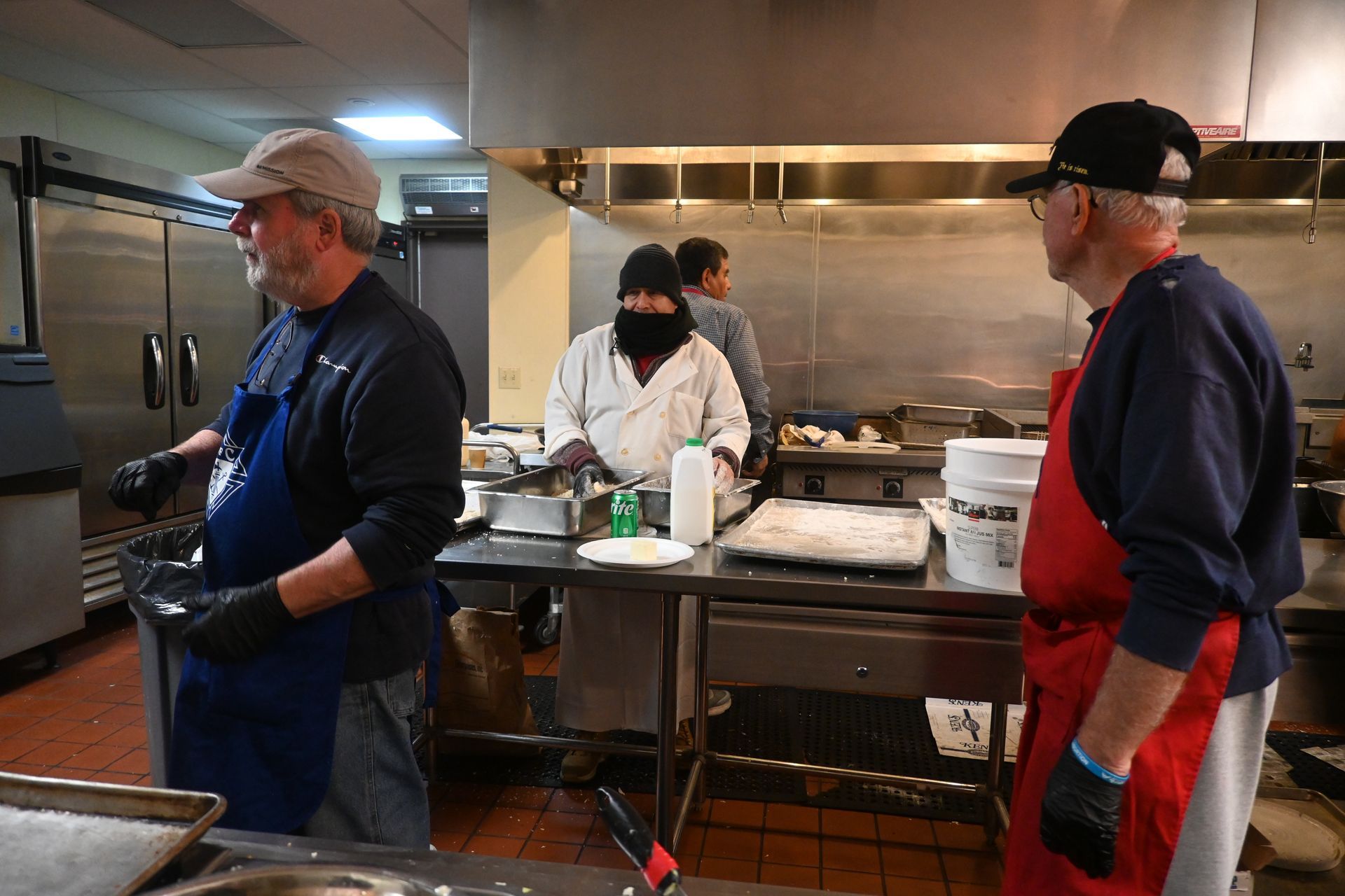 People in aprons working around a stainless-steel kitchen prep table with trays and cookware