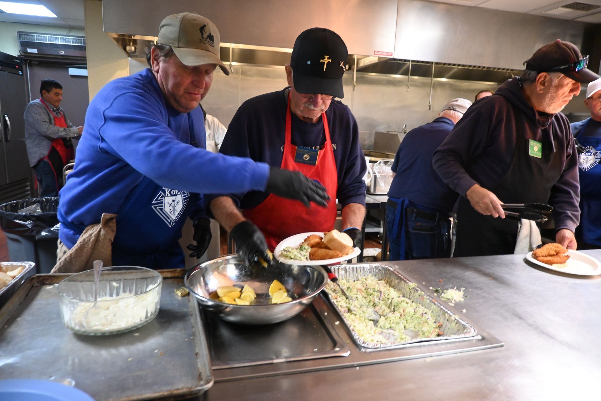 Volunteers serving food from a buffet line in a kitchen, with trays and a white plate in front.