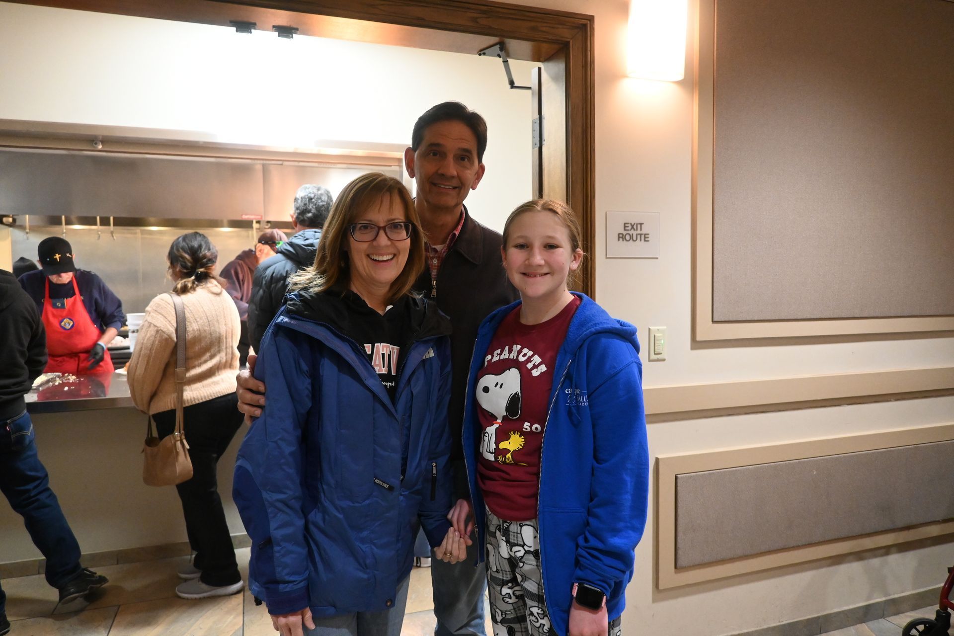 Three people smiling in blue jackets standing together indoors near a mirror and wall decor