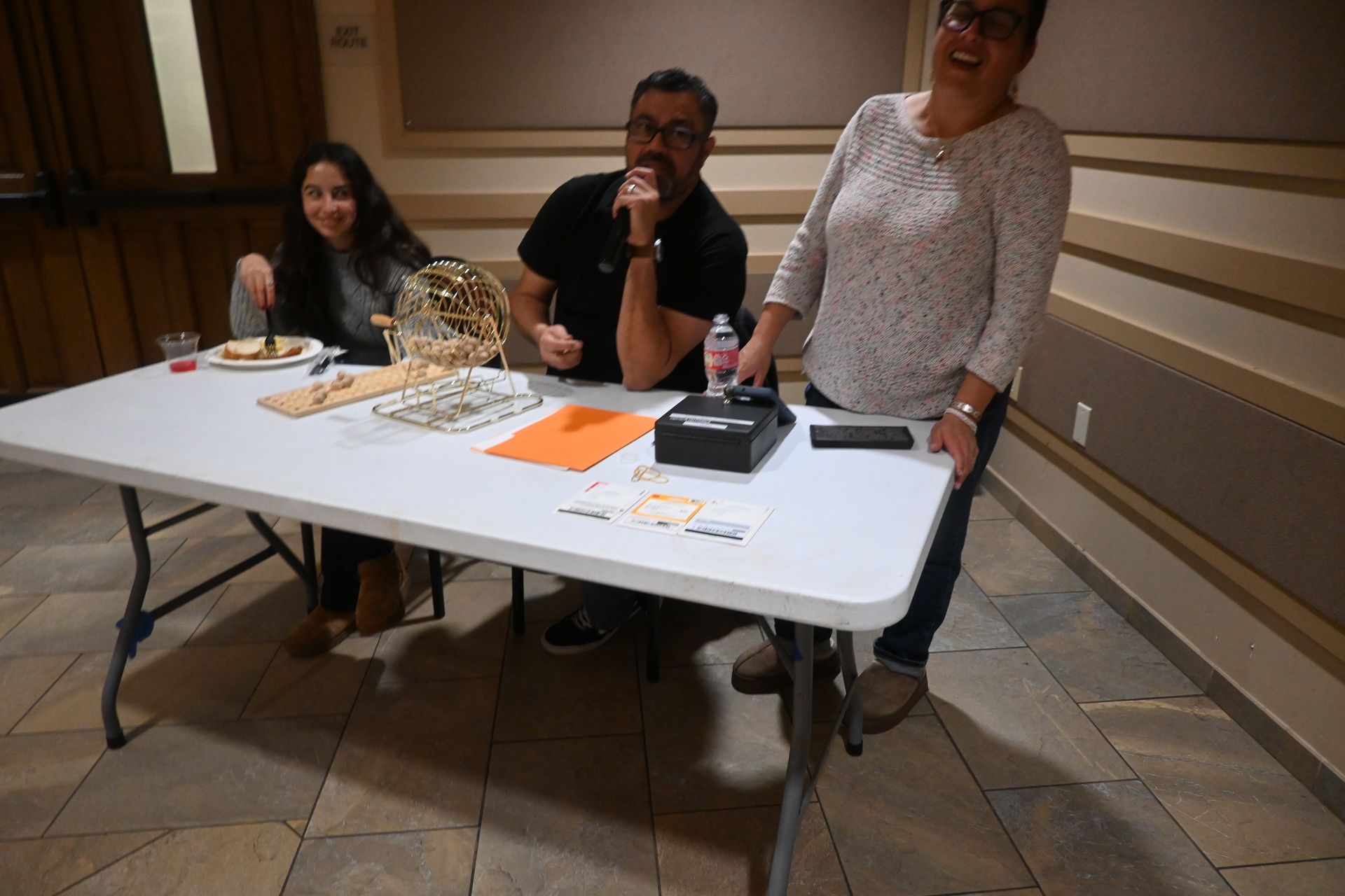Three people seated at a table with papers and snacks in a meeting room, one person standing and smiling.