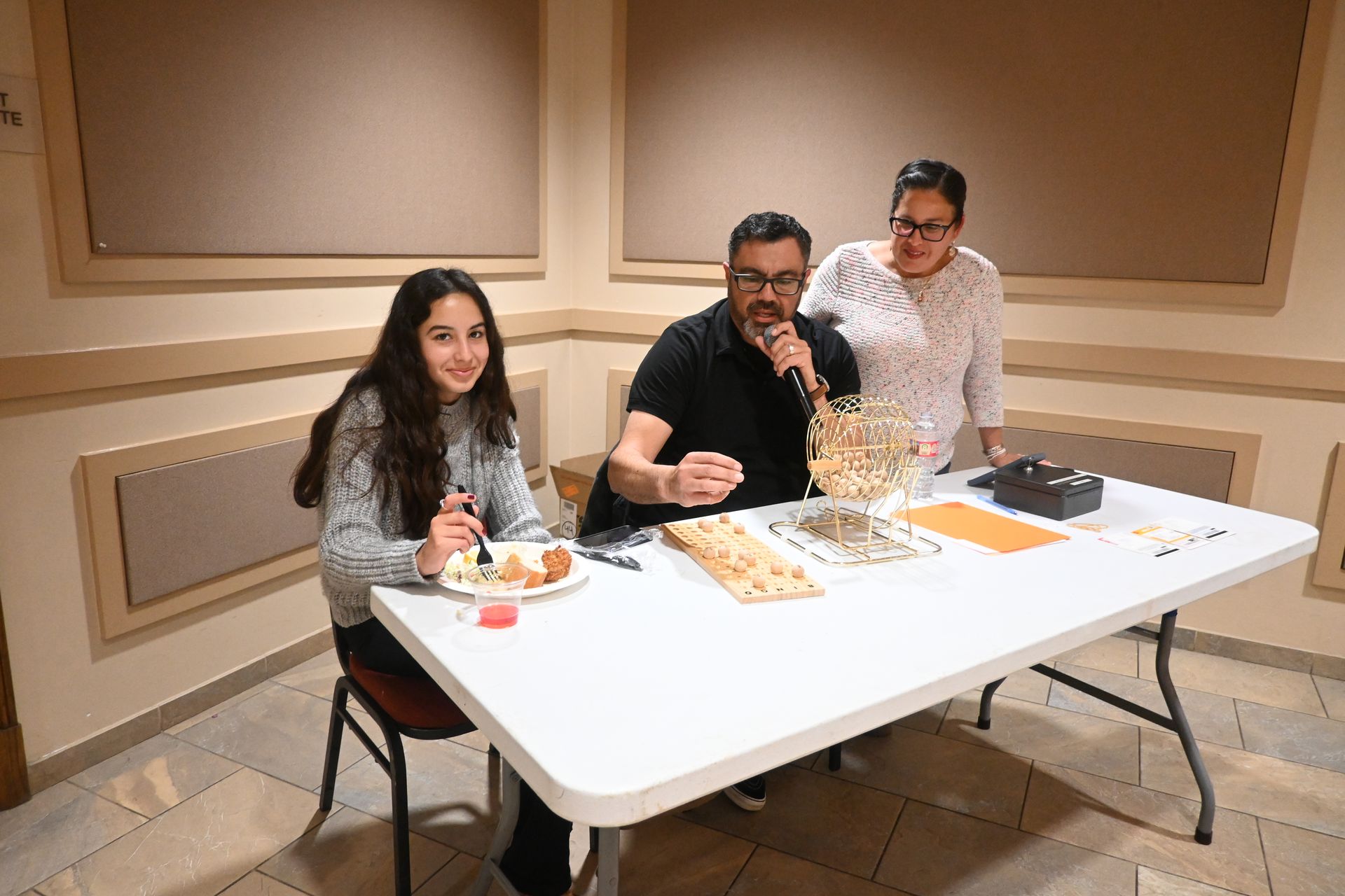 Three people seated at a table in a beige room, sharing food and playing a board game.