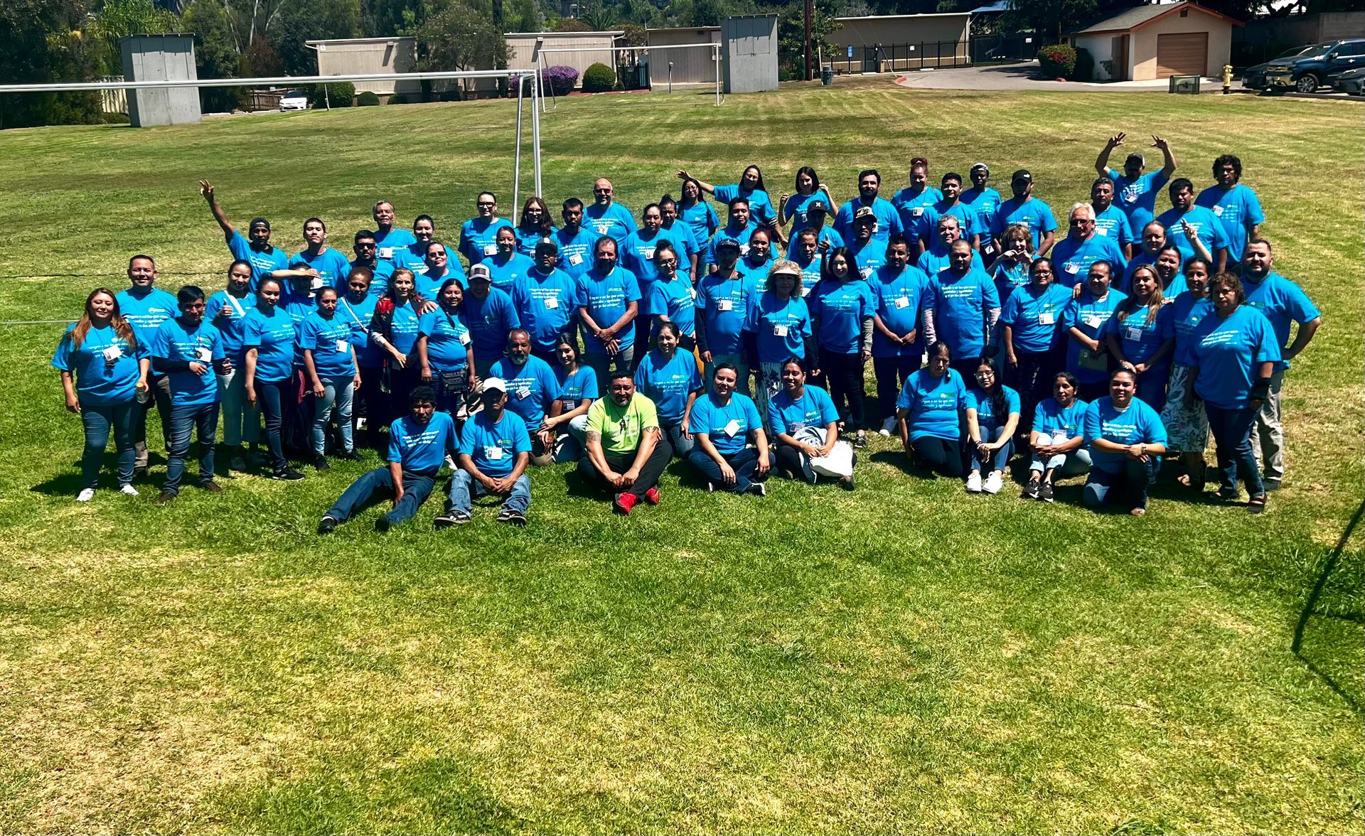 Large group in matching blue shirts posing outdoors on a sunny grassy field