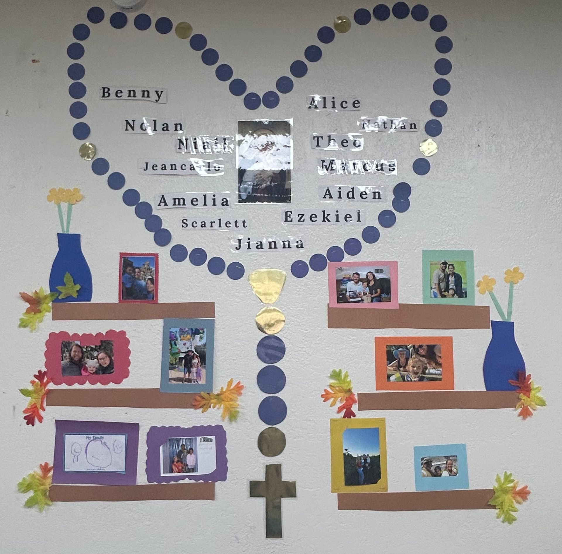 Heart-shaped memorial board with photos, names, blue dots, and floral decorations on a wall.