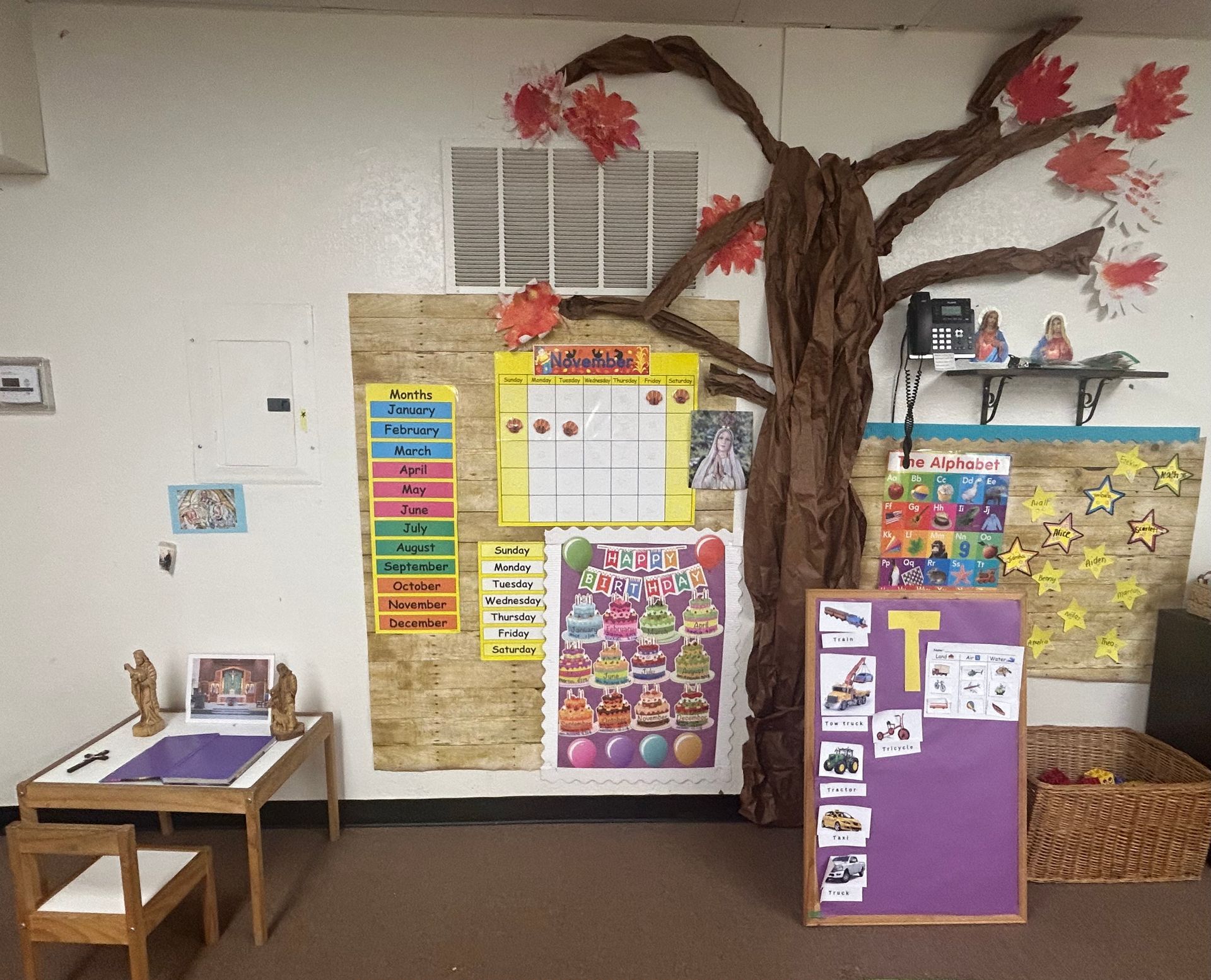 Classroom wall with colorful bulletin boards and a large tree mural beside a small desk