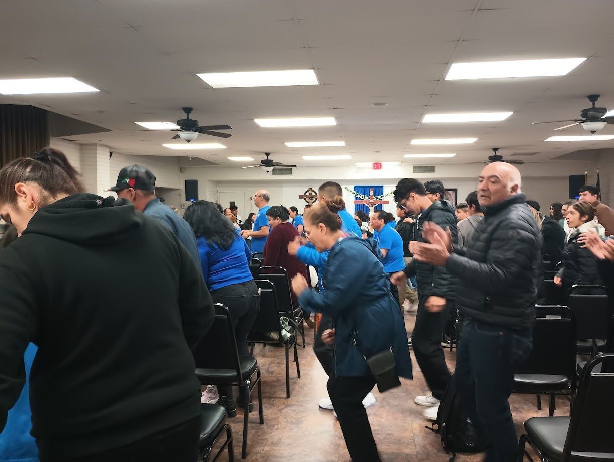 Crowded indoor event room with people standing and clapping near tables under bright fluorescent lights