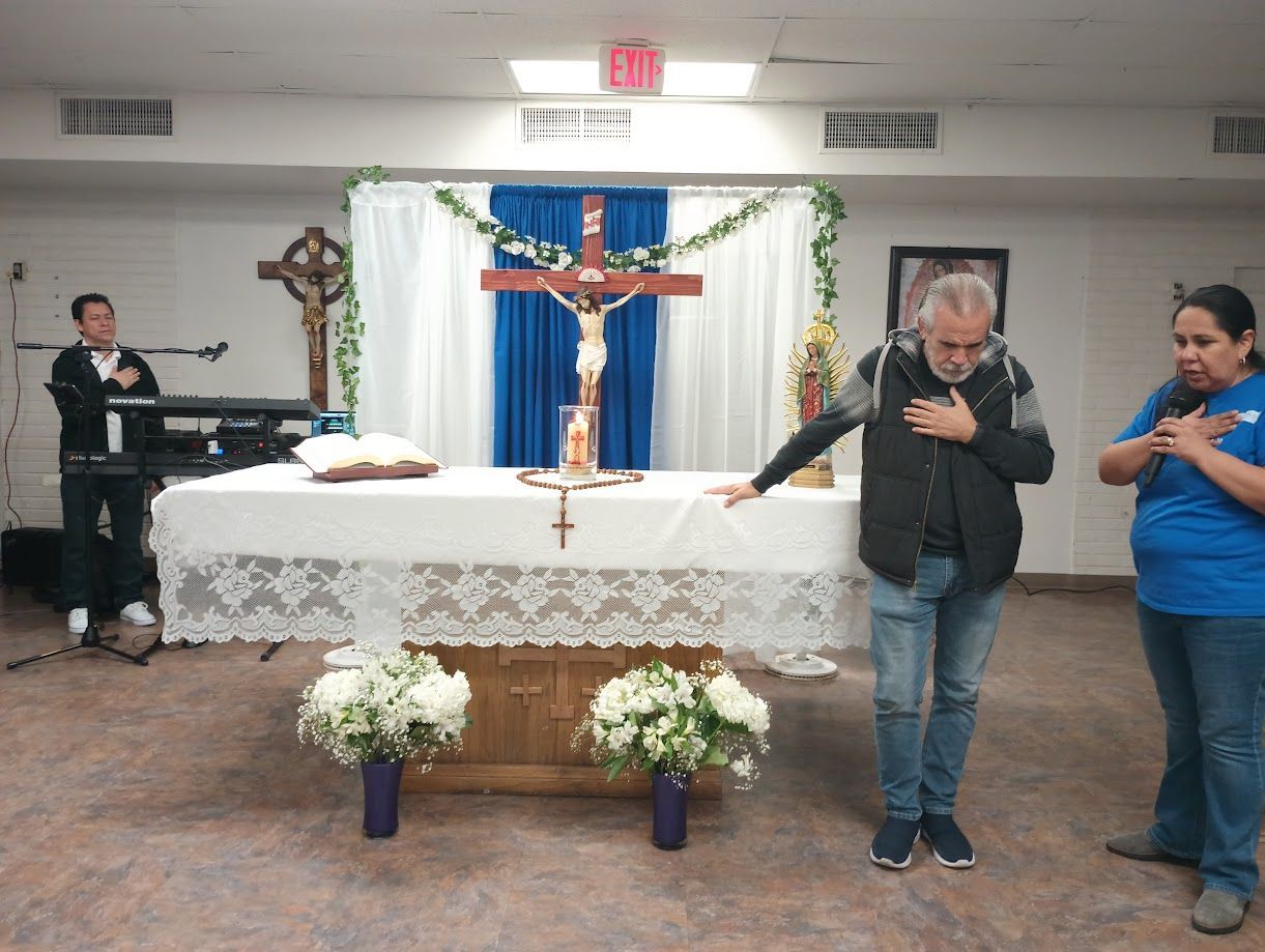Church altar with crucifix and flowers; two people stand praying in the foreground.