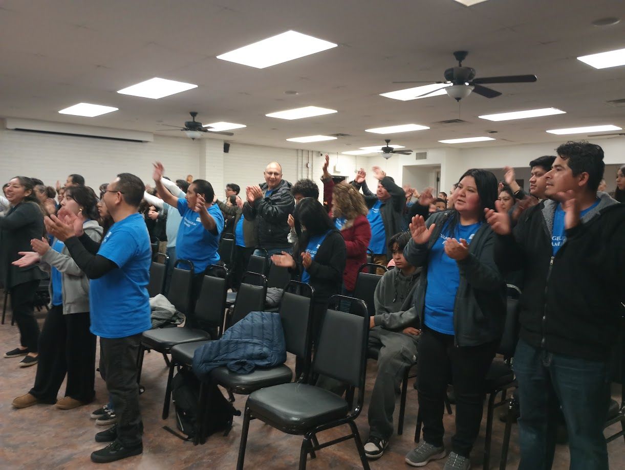 People in blue shirts standing and clapping in a crowded meeting room.